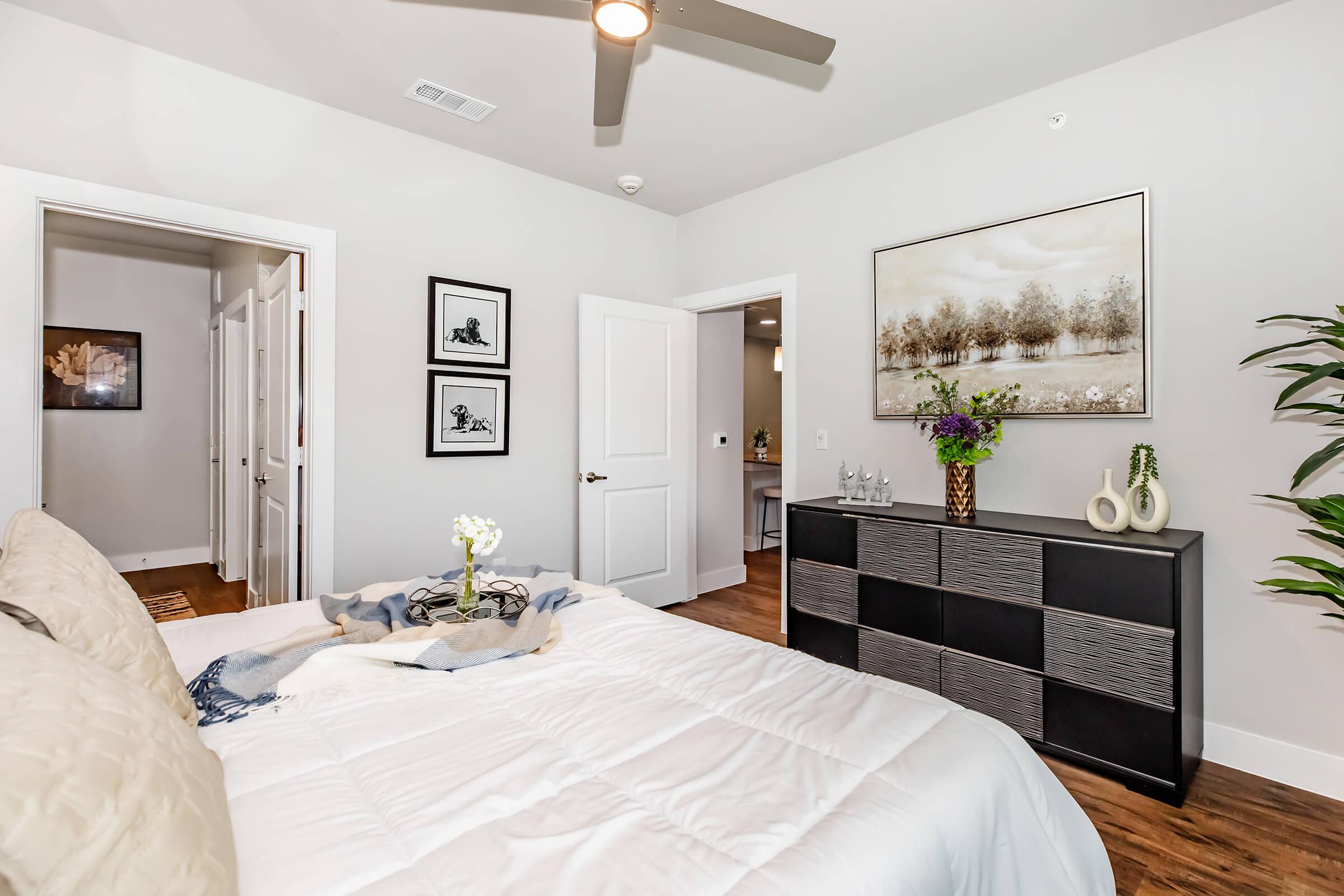 A cozy bedroom featuring a neatly made bed with a light-colored duvet, decorative pillows, and a throw blanket. The room has a modern look with framed artwork on the walls, a potted plant, and a stylish black dresser. Light streams in through the doorways, enhancing the inviting atmosphere.