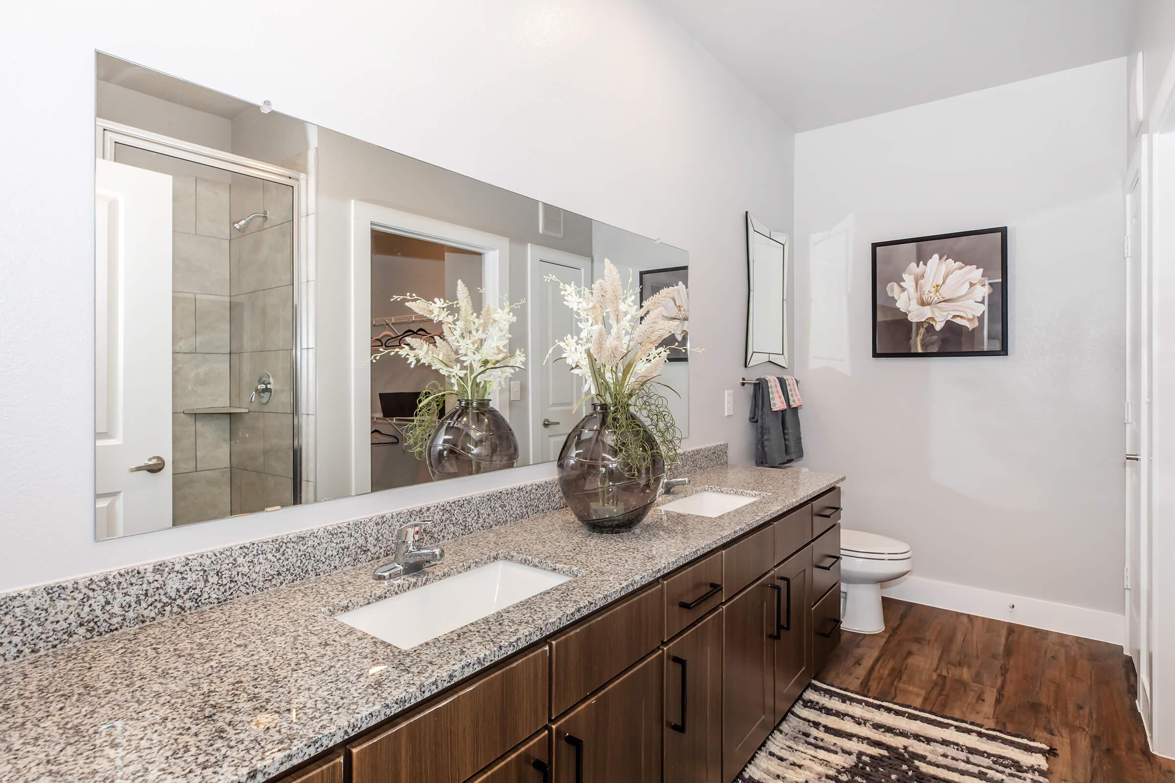 Modern bathroom featuring a double vanity with a granite countertop, two sinks, and a large mirror. Decorative floral arrangements in vases sit atop the counter. Wall art is visible, and a shower is seen in the background behind a glass door. The floor has wooden planks, and there is a toilet in the corner.