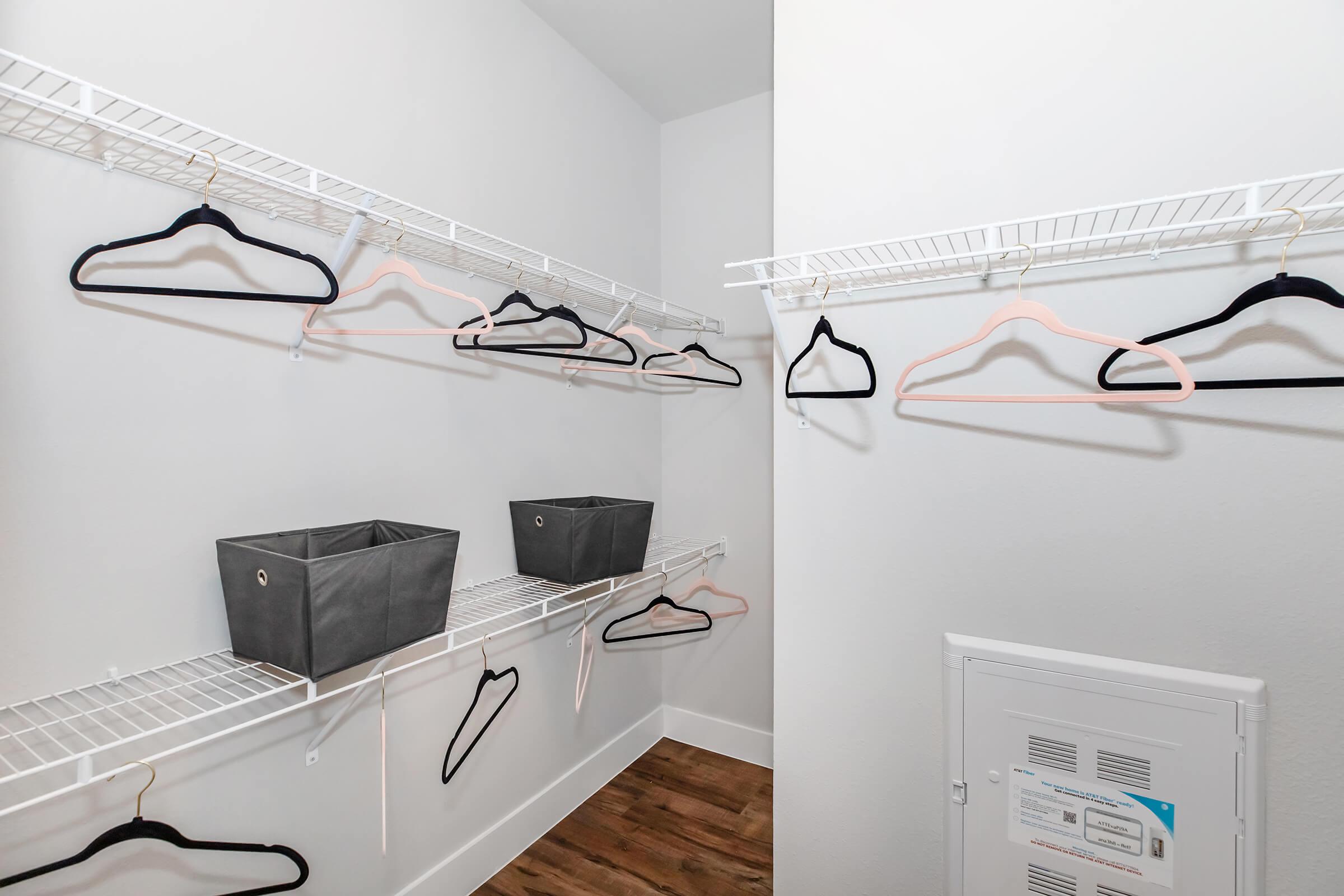 A walk-in closet featuring metal wire shelving with several empty black and pink hangers. Two gray storage bins are placed on the shelving, and the floor has wooden laminate. A small white electrical panel is visible on the wall. The overall decor is minimalist and clean.