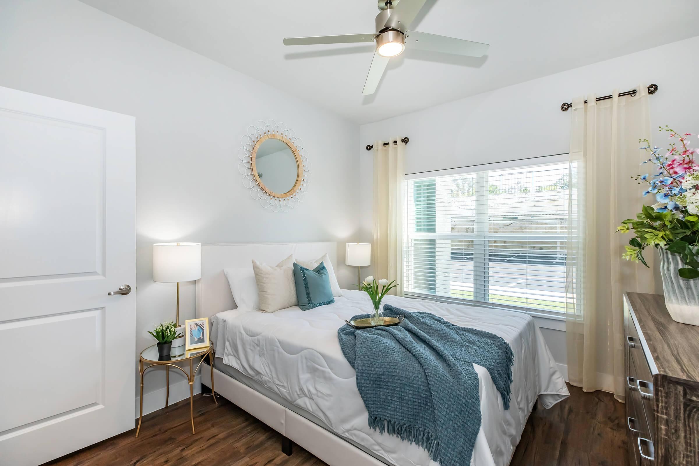A bright, modern bedroom featuring a queen-sized bed with white bedding and decorative pillows. A light blue throw is draped over the bed. There are two bedside lamps on gold side tables, a circular mirror on the wall, and a vase with flowers on a dresser. Soft natural light enters through the window.