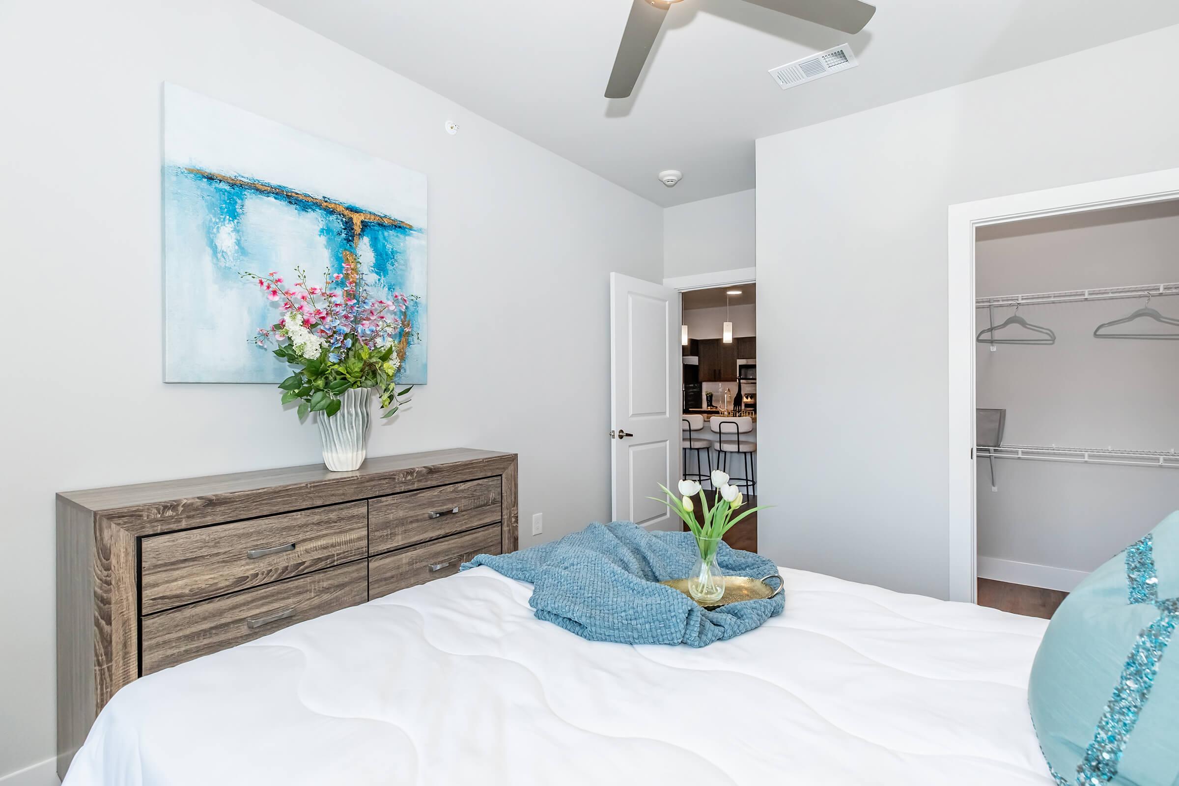 A bright bedroom featuring a bed with a white quilt, a wooden dresser with a decorative vase filled with flowers, and a cozy blue throw. The walls are painted light gray, and there's a painting above the dresser. A doorway in the background leads to a kitchen area, with a closet to the side.