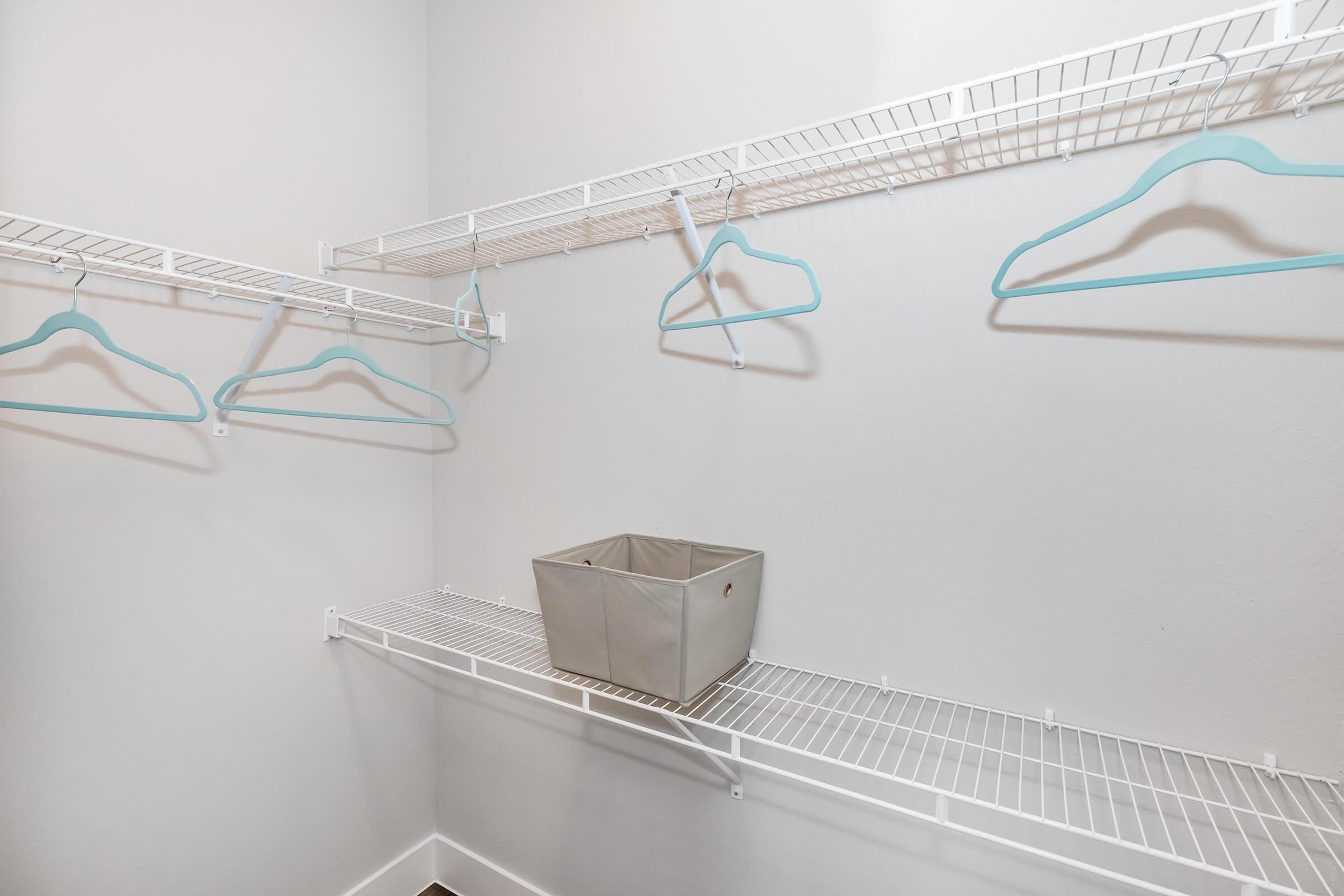 A neatly organized closet featuring white wire shelving. There are several light blue hangers hanging on the shelves, and a gray storage basket sits on the lower shelf. The walls are painted light gray, creating a clean and spacious appearance.