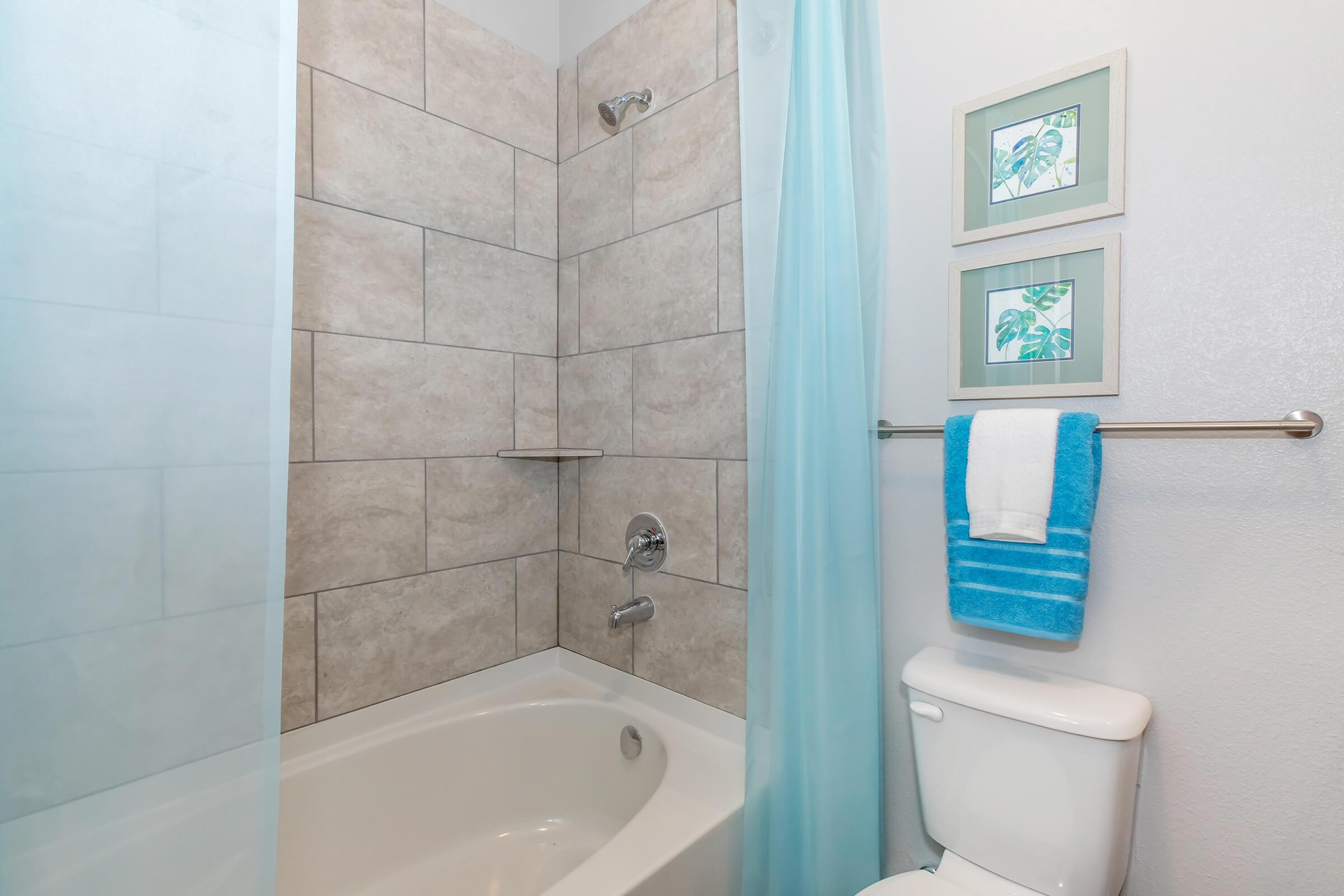 A modern bathroom featuring a bathtub with a shower, light-colored tiled walls, a blue shower curtain, and two framed artworks above a small shelf. A white toilet is positioned beside a towel rack holding a blue towel and a white towel.
