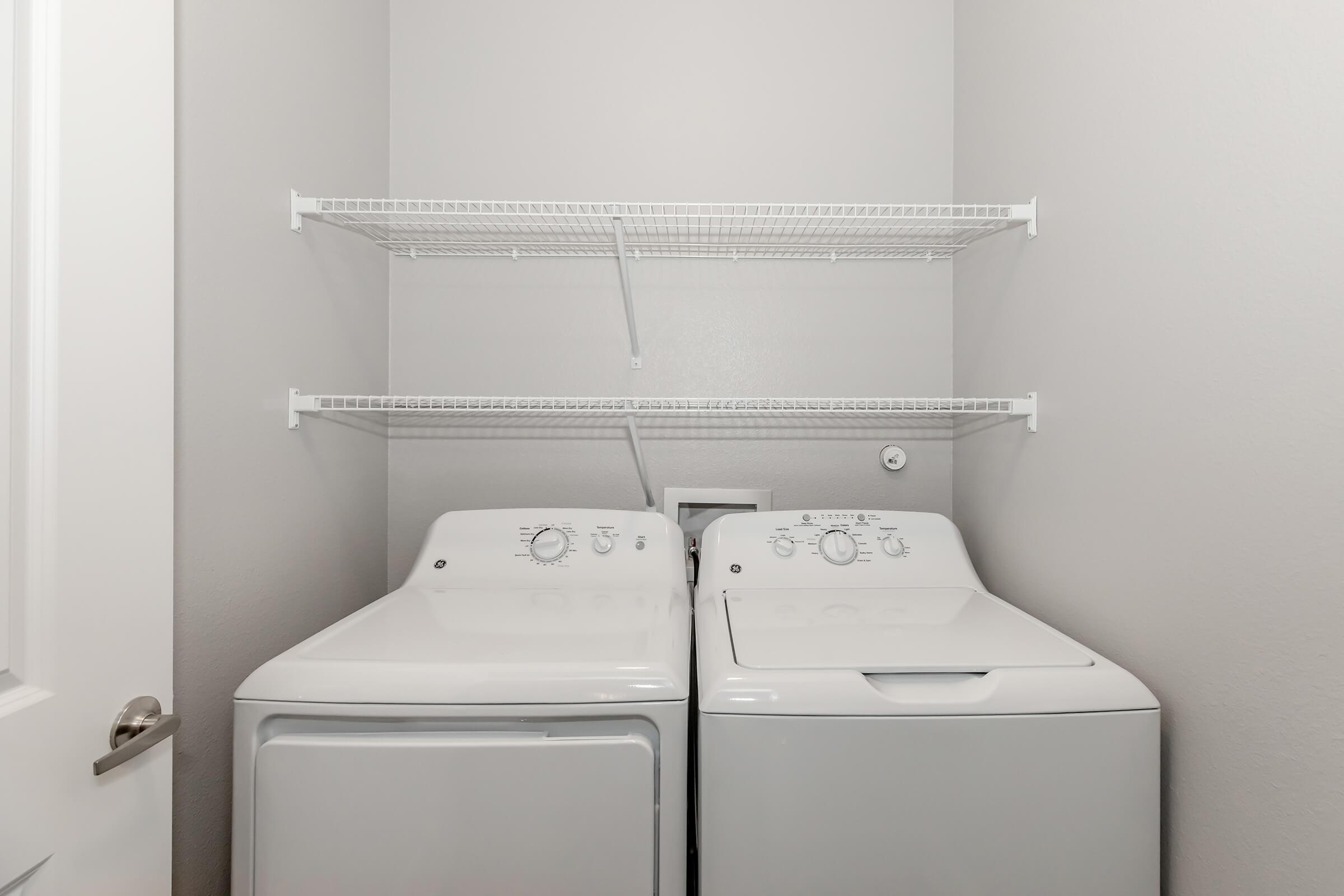 A laundry room featuring a white washer and dryer side by side against a light gray wall. Above them, there are two wire shelves for storage. A door is partially visible on the left side of the image.