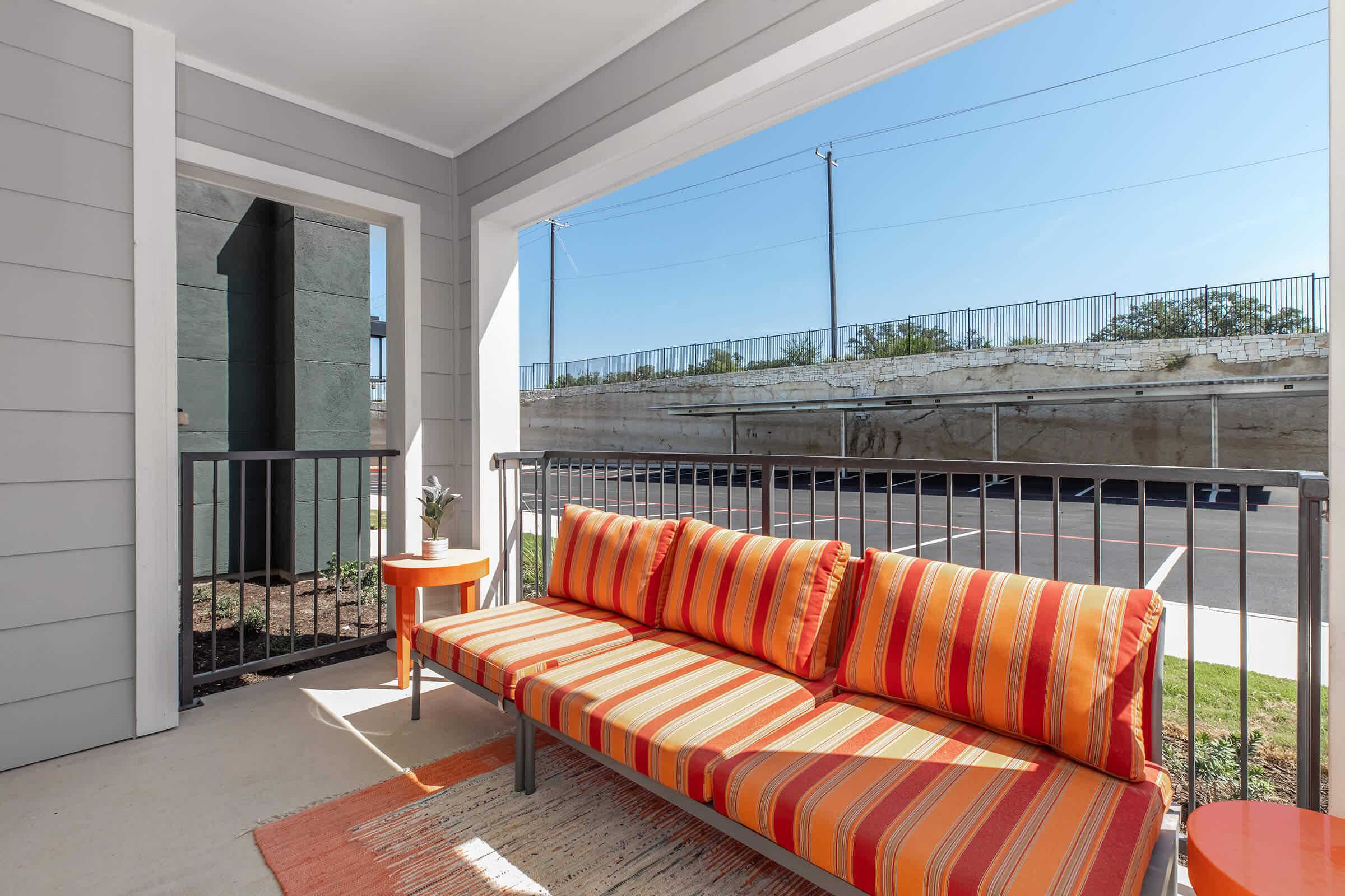 A brightly colored outdoor patio area featuring a striped orange and red couch, a small round orange table, and a modern railing. The backdrop includes a clear blue sky and a concrete structure in the distance. The setting conveys a cozy and inviting atmosphere for relaxation.
