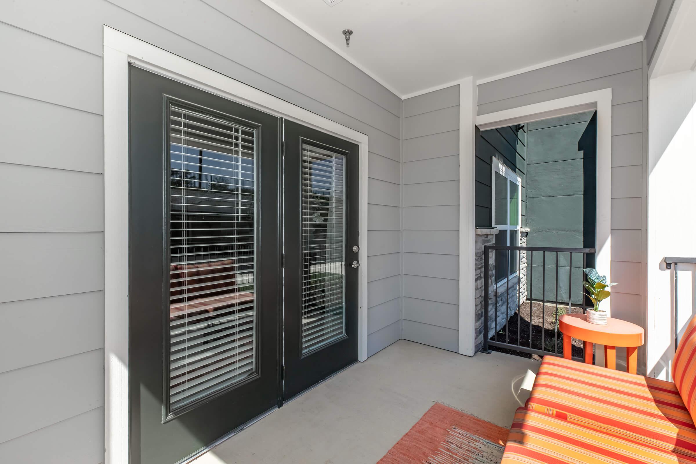 A corner of a balcony featuring a pair of glass-paneled double doors with blinds, leading to an indoor space. The balcony has a wooden railing and a small orange-striped chair placed against a gray wall. The scene is well-lit and shows a blend of indoor and outdoor living.