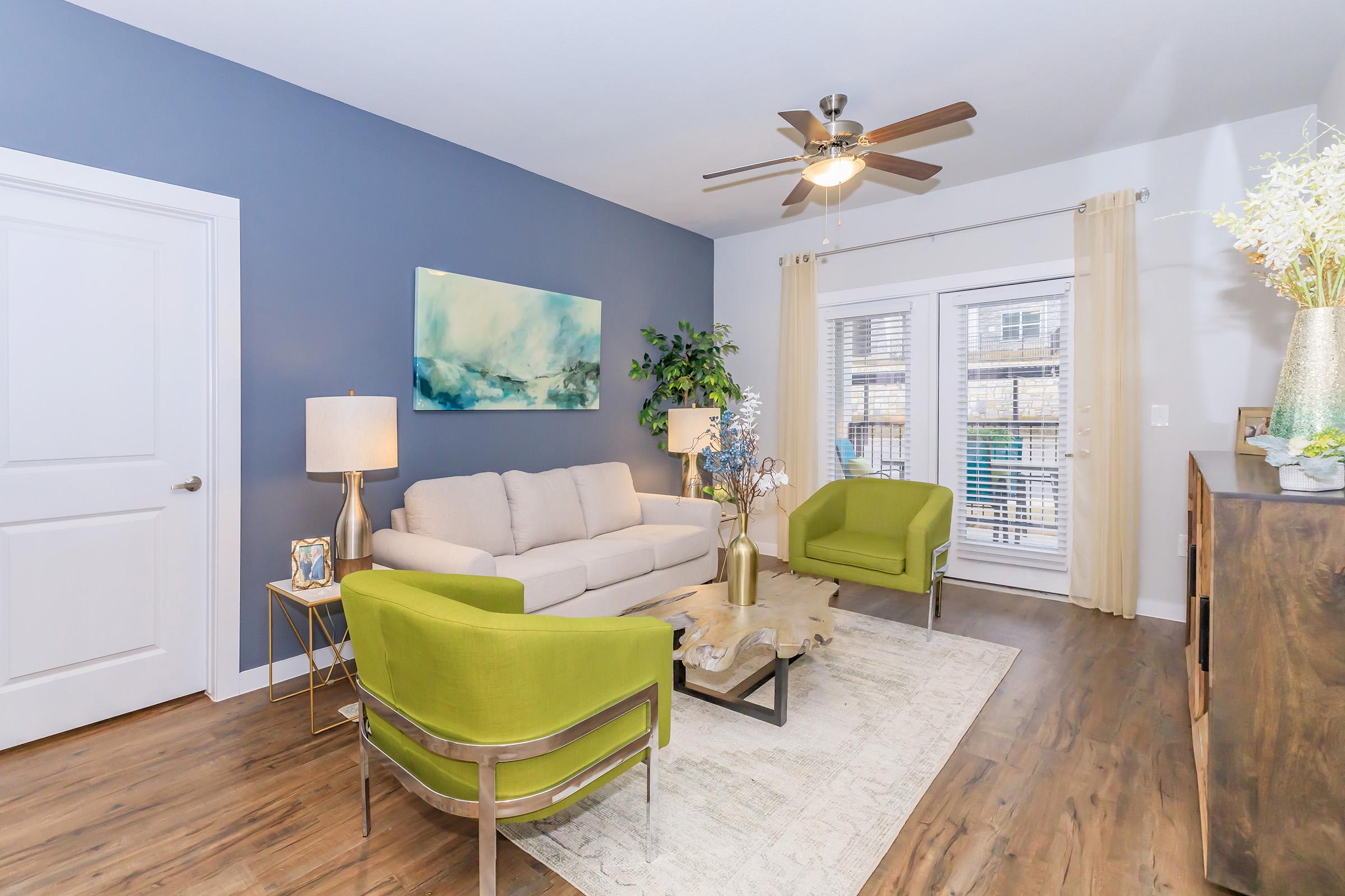 A modern living room featuring a light gray sofa, two vibrant green chairs, and a wooden coffee table. There is a ceiling fan above, a floor lamp beside the sofa, and a large piece of abstract art on the wall. Bright curtains frame a window that opens to a view outside. The decor is stylish and inviting.