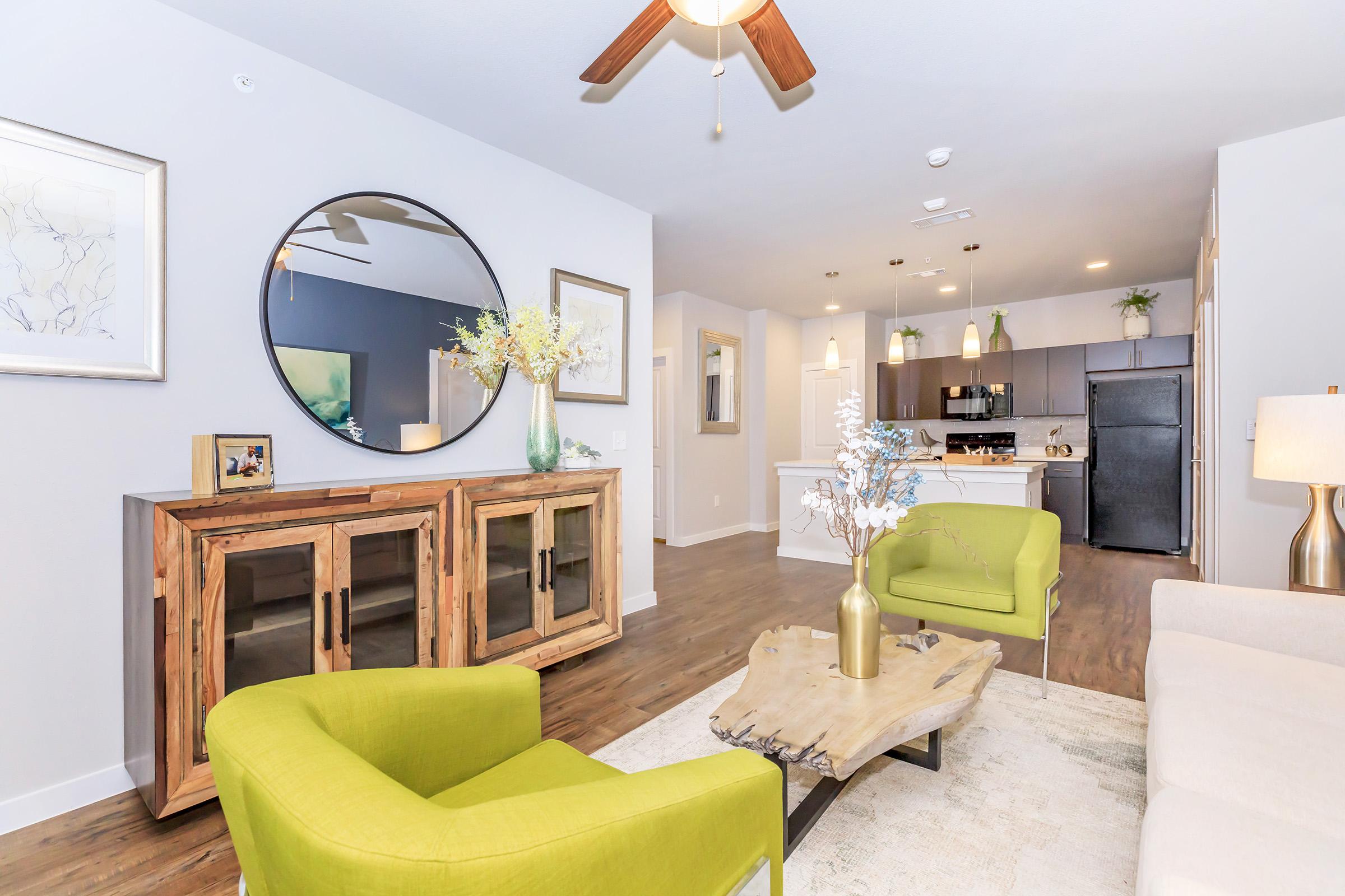 Interior of a modern living space featuring a green armchair, a light sofa, and a wooden coffee table. There's a decorative circular mirror on the wall and a wooden cabinet. Open kitchen visible in the background with black appliances and light-colored cabinetry. Natural light brightens the room.