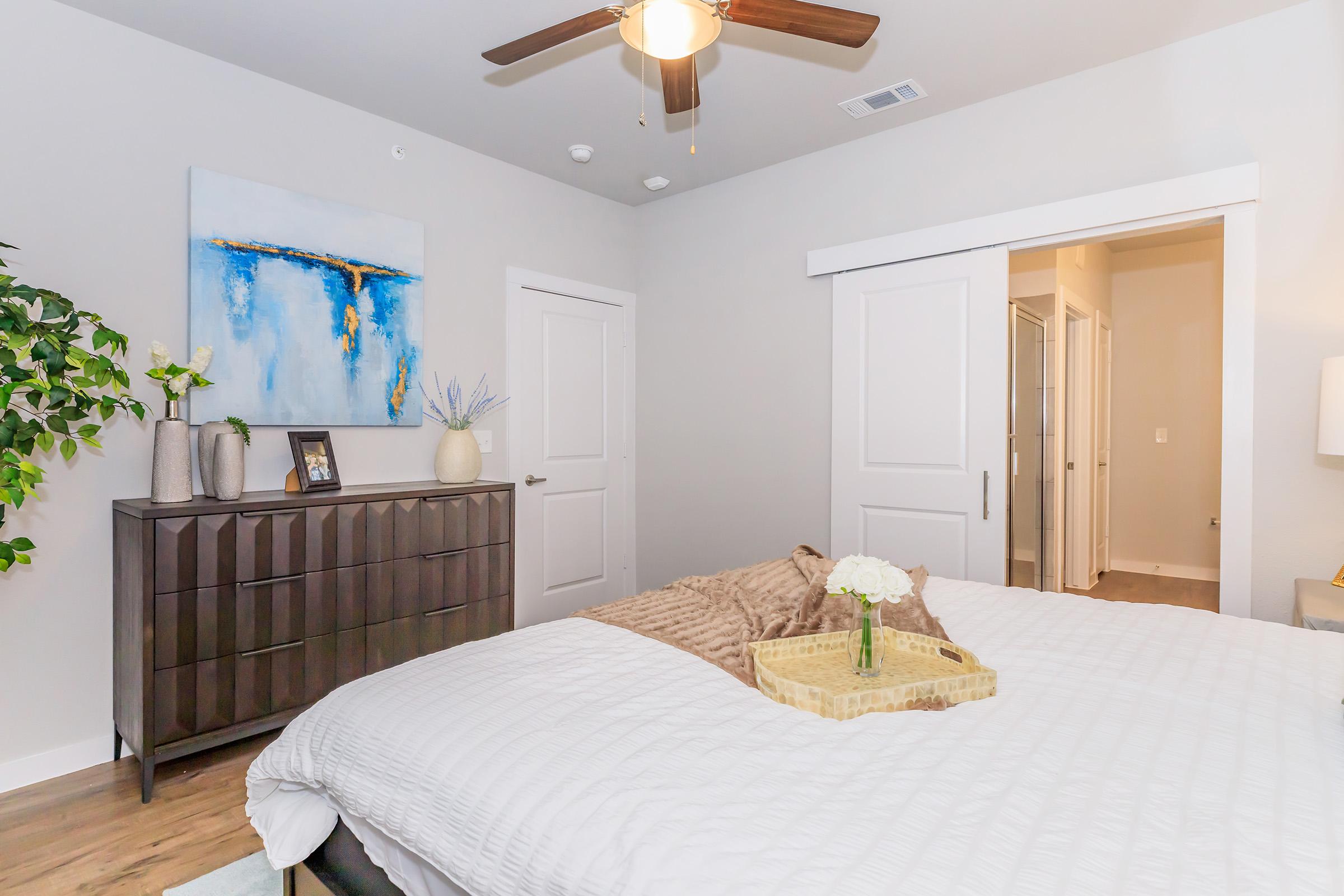 A cozy bedroom featuring a neatly made bed with a light-colored comforter and a decorative throw. A wooden dresser is positioned against the wall, complemented by a potted plant. Artwork hangs above the dresser, and natural light fills the space from a window behind a door. A ceiling fan adds a modern touch.