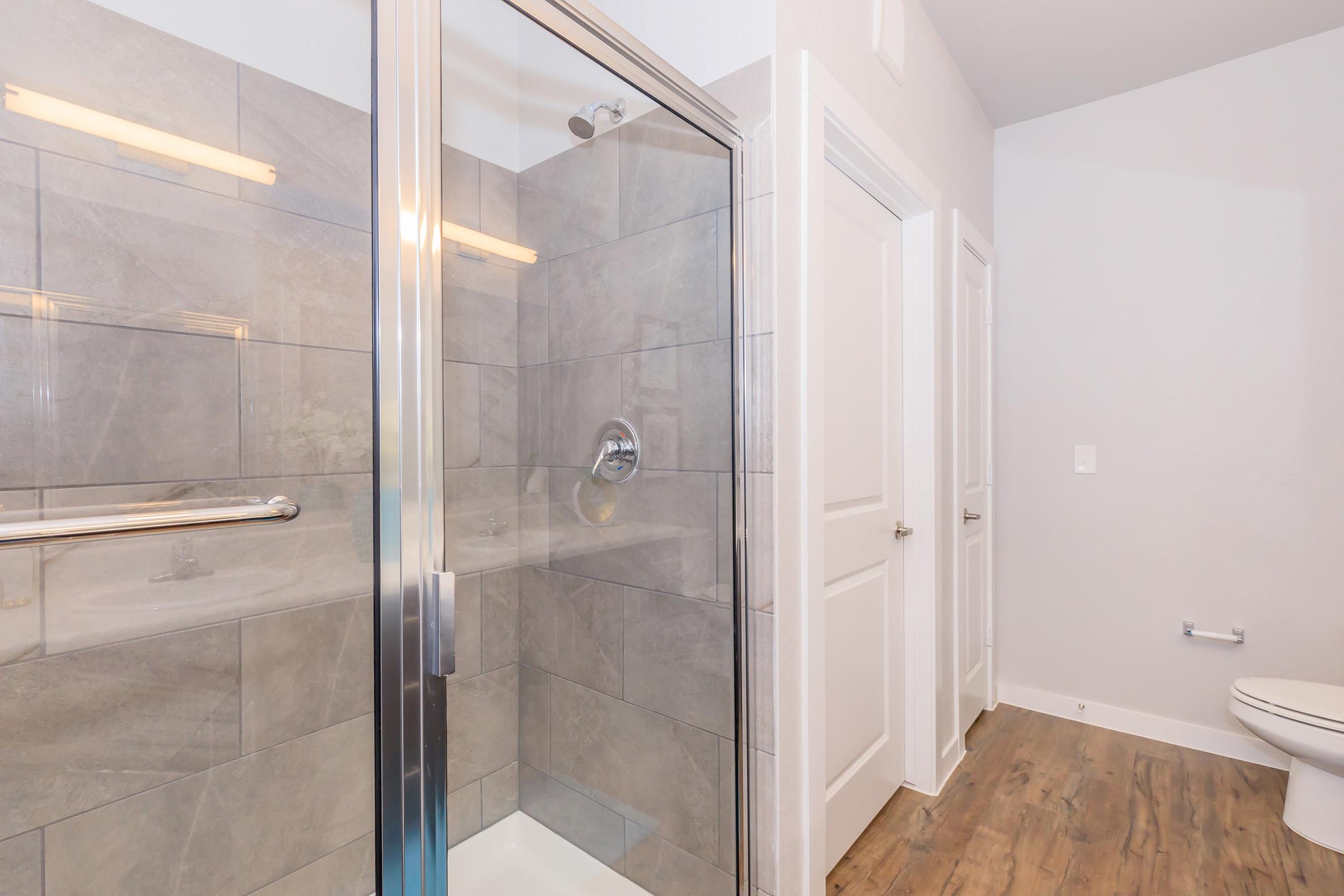 A well-lit modern bathroom featuring a glass shower enclosure with gray tiled walls, a showerhead, and a sliding door. There are two white doors leading to separate areas, and a toilet is visible in the corner. The flooring is wood-like, adding a warm touch to the space.
