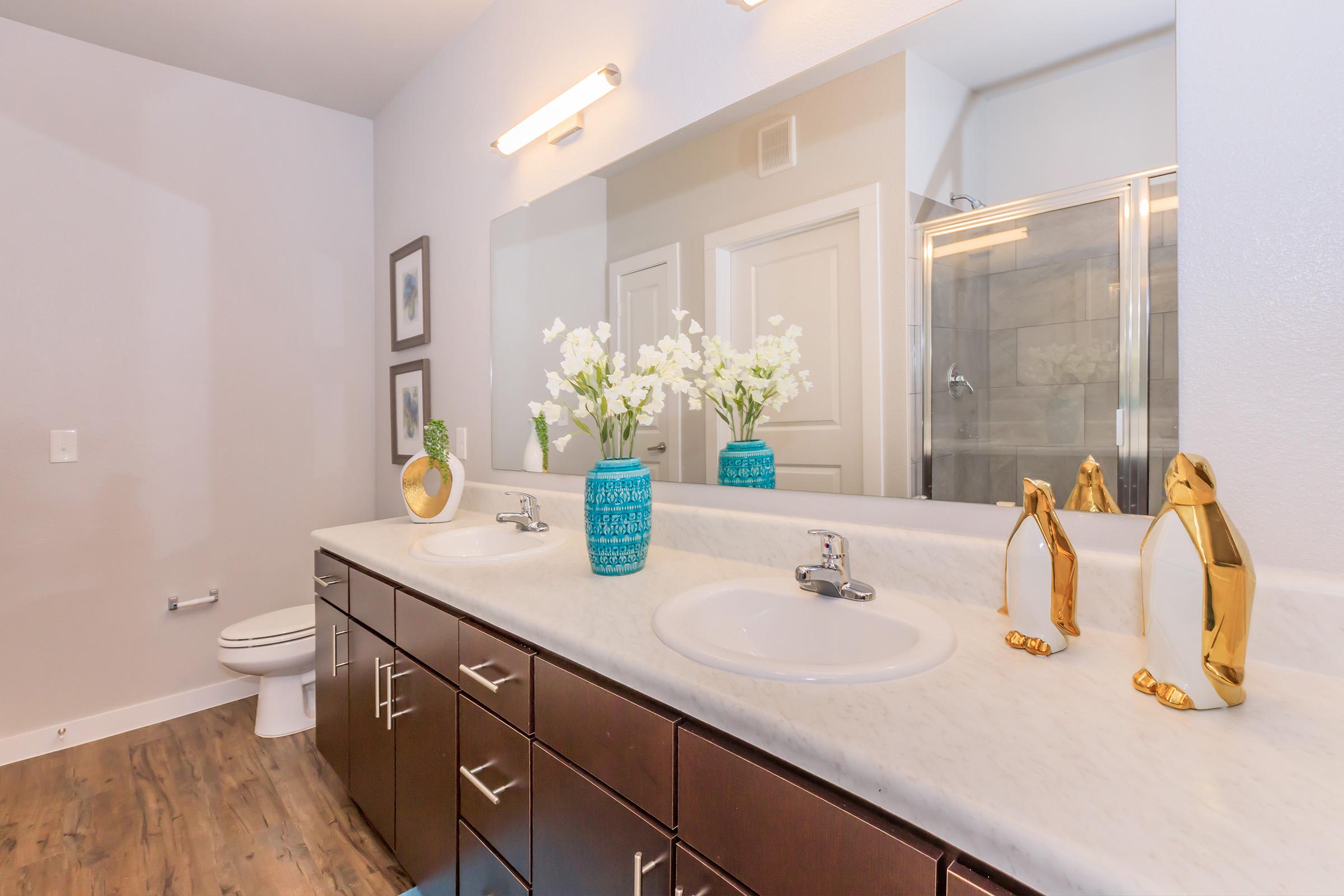 A modern bathroom featuring a double vanity with a light-colored countertop, two sinks, and stylish faucets. The dark wooden cabinetry contrasts with the countertop. Above the vanity, there's a large mirror and decorative elements including a blue vase with white flowers. A shower area is visible in the background.