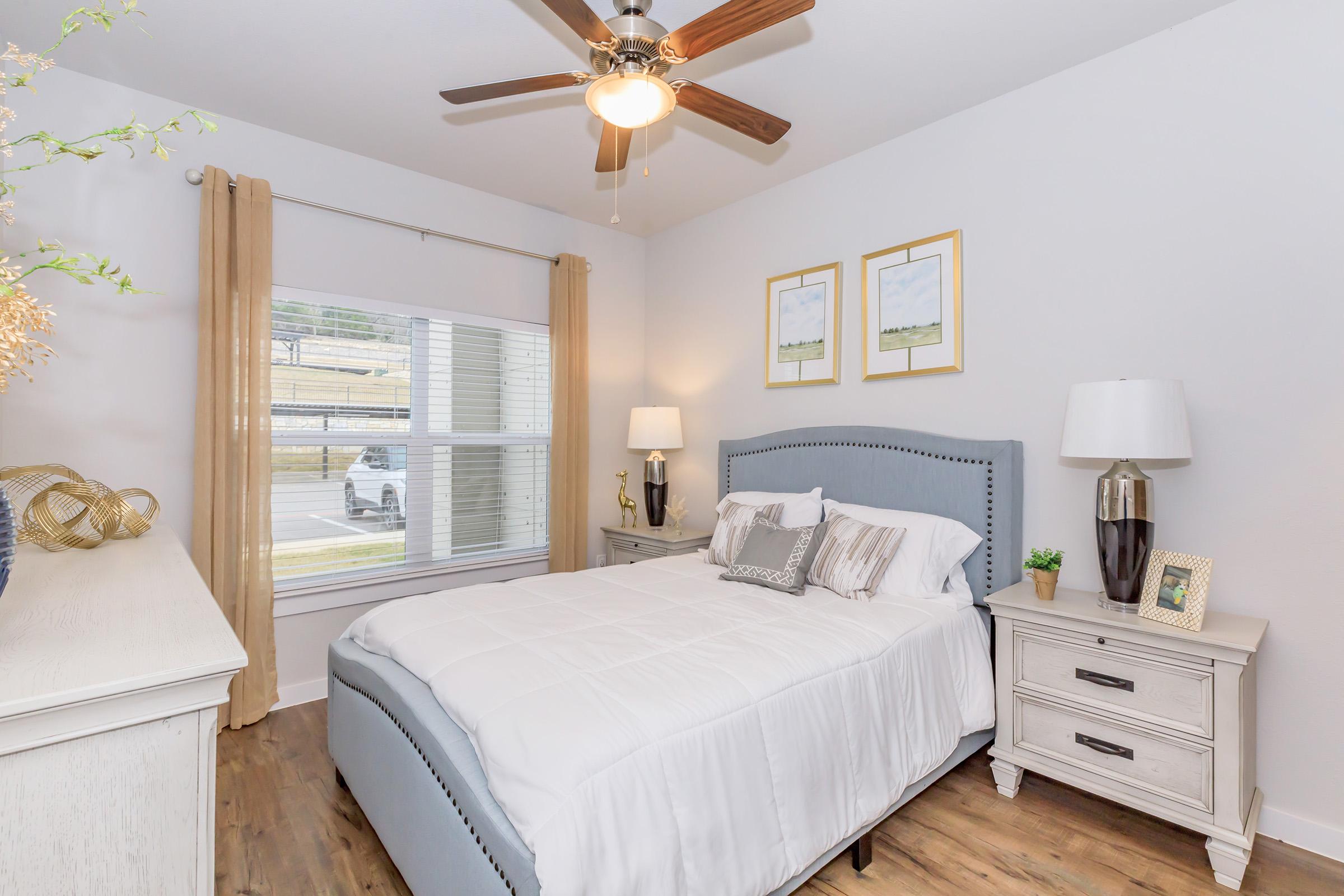 A cozy bedroom featuring a light blue upholstered bed with decorative pillows, a wooden nightstand with a lamp and small plant, a dresser, and framed artwork on the walls. Natural light comes through a window with sheer curtains, complemented by a ceiling fan. Hardwood flooring adds warmth to the space.