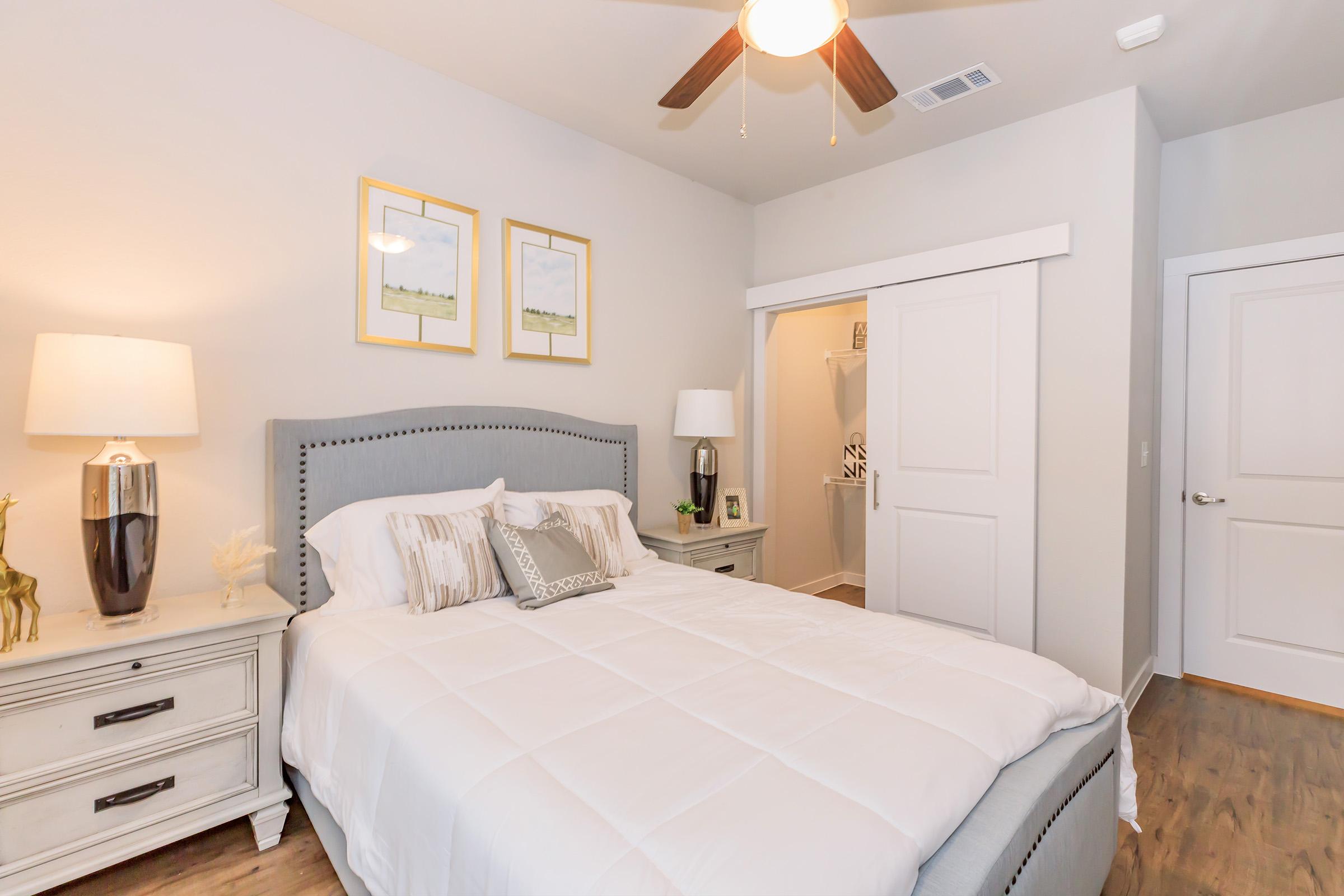 A cozy bedroom featuring a light gray upholstered bed with decorative pillows. There are two framed artworks above the bed, a bedside table with a lamp, and another table with a decorative item. A white closet door is visible, along with a ceiling fan and warm wood flooring, creating a welcoming atmosphere.