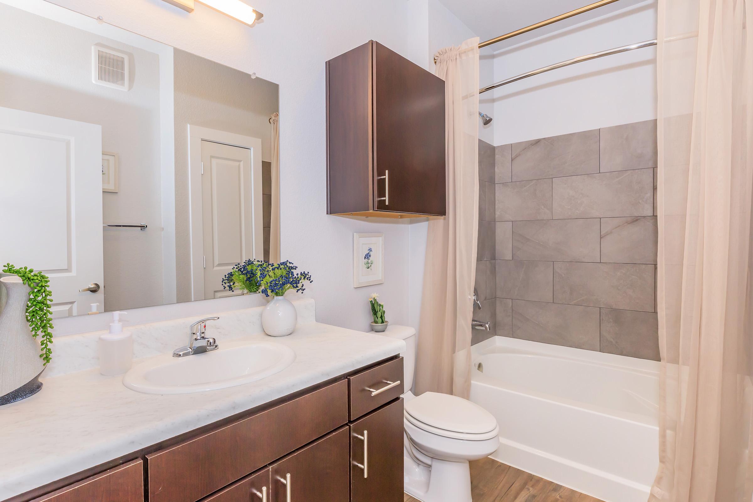 A modern bathroom featuring a light-colored marble countertop with a sink, a large mirror above the sink, dark wood cabinetry, and a shower-tub combination with gray tiles. Decorative plants are present on the counter, and a cream-colored shower curtain hangs by the tub.