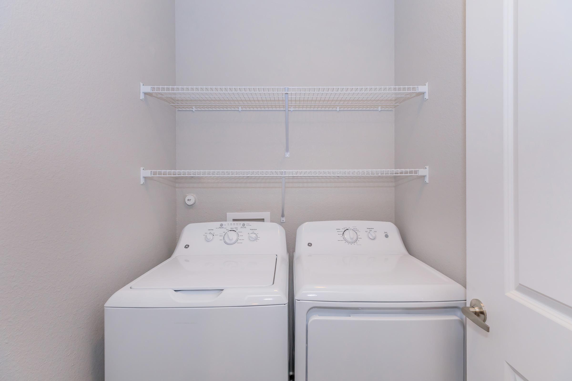 A laundry room featuring a white washing machine and a dryer side by side, with wire shelving above for storage. The walls are painted a light color, and the room has a door that is partially open, suggesting an organized and functional laundry space.
