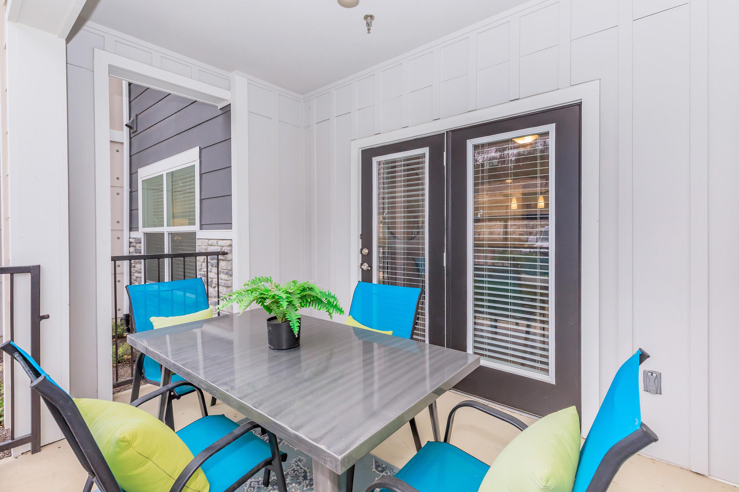 A modern porch area featuring a gray table surrounded by bright blue chairs with green cushions. A small potted plant sits in the center of the table. Sliding glass doors are visible in the background, leading to an indoor space, with light-colored wood paneling and dark exterior walls.