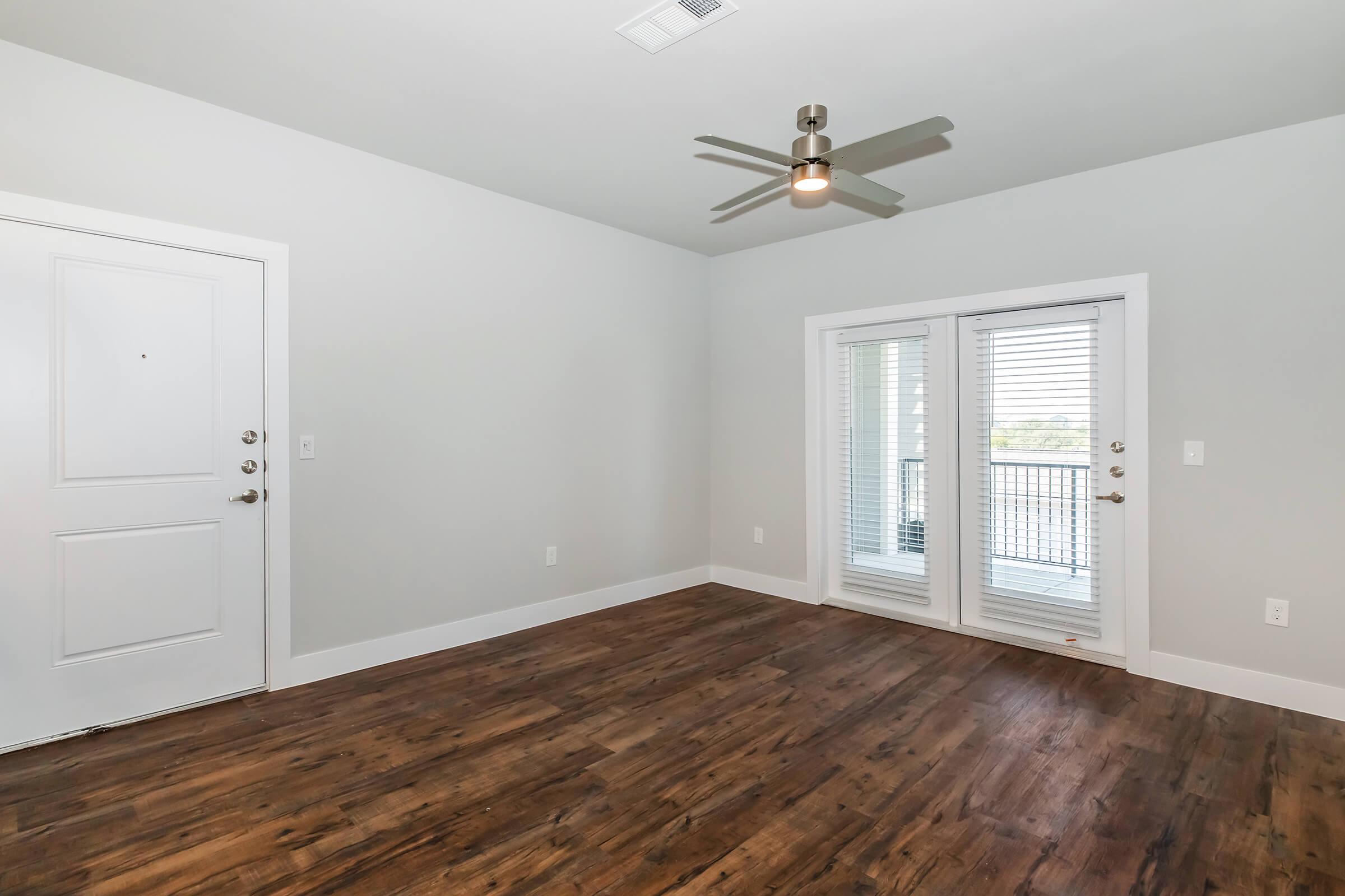 A spacious, empty living room with light gray walls and wood-style flooring. A ceiling fan is installed, and there are two large sliding glass doors with white blinds that lead to a balcony. The front door is visible on the left side of the image. The room is well-lit with natural light.
