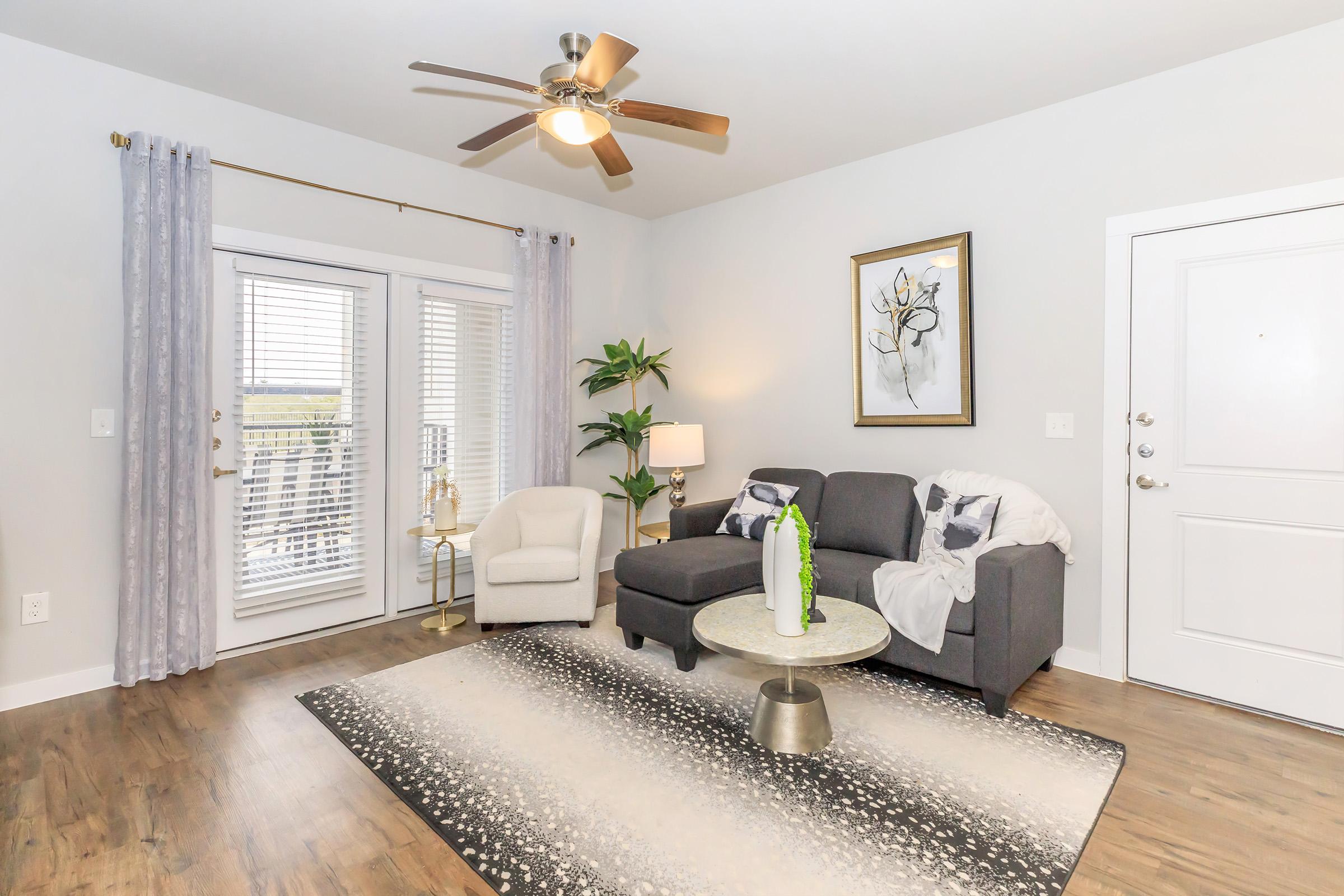 A stylish living room featuring a gray sectional sofa, a round coffee table, and a light-colored accent chair. The space has large windows with blinds, a ceiling fan, and a decorative plant. A modern artwork hangs on the wall, and a plush area rug is underfoot, creating a cozy and inviting atmosphere.