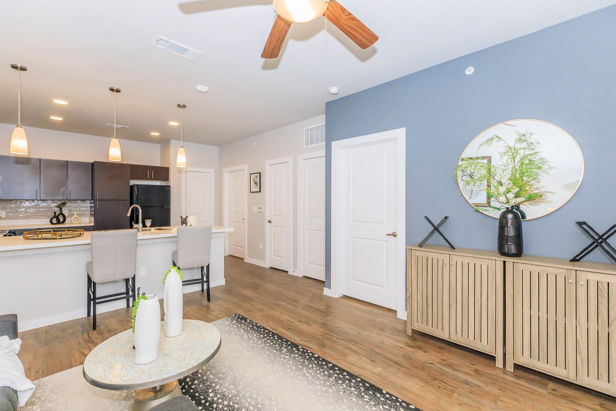 A modern living space featuring an open kitchen with dark cabinets, a granite countertop, and bar stools. The living area includes a decorative table with vases, a stylish rug, and a console table against a blue accent wall. Soft lighting from pendant fixtures complements the welcoming atmosphere.