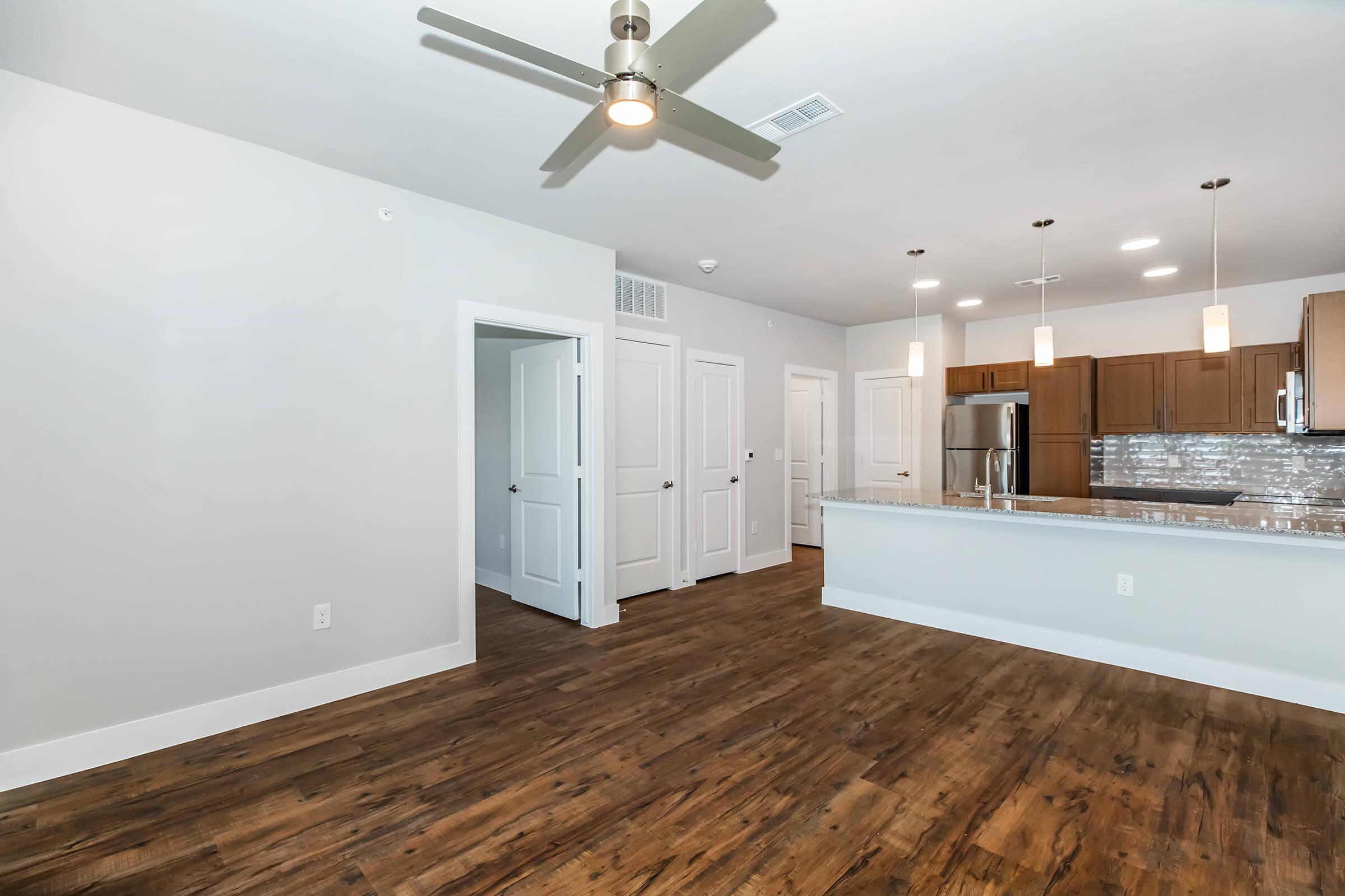 Modern living room with wooden flooring and neutral-colored walls. Features a ceiling fan, a spacious kitchen area with sleek cabinets and counter space, stainless steel appliances, and pendant lighting. A doorway leads to another room, creating an open and inviting atmosphere.