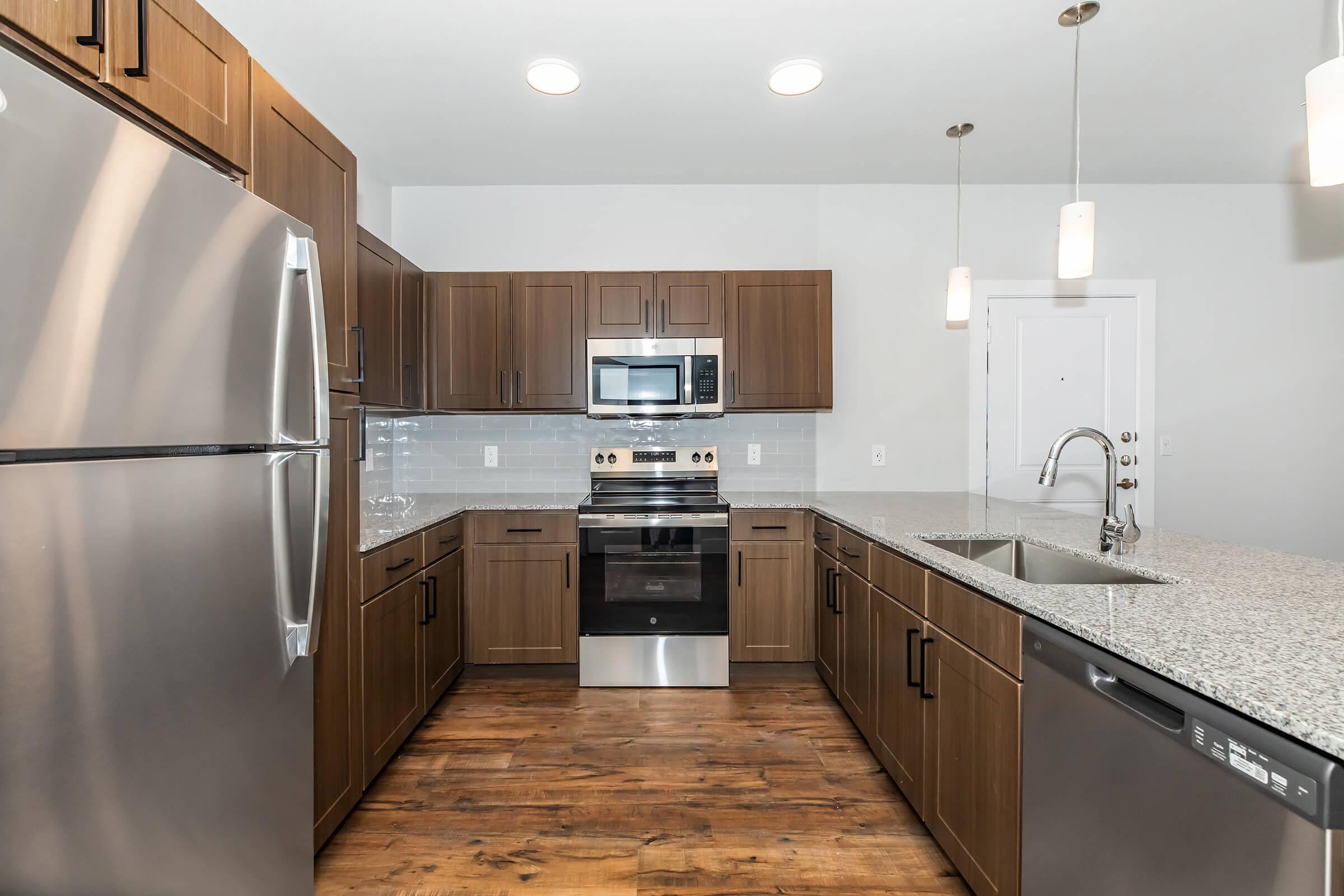Modern kitchen featuring wooden cabinets, stainless steel appliances including a refrigerator, microwave, and oven, a granite countertop, and a dual-basin sink. The room is well-lit with recessed ceiling lights and pendant lighting above the countertop, set against a neutral wall color.