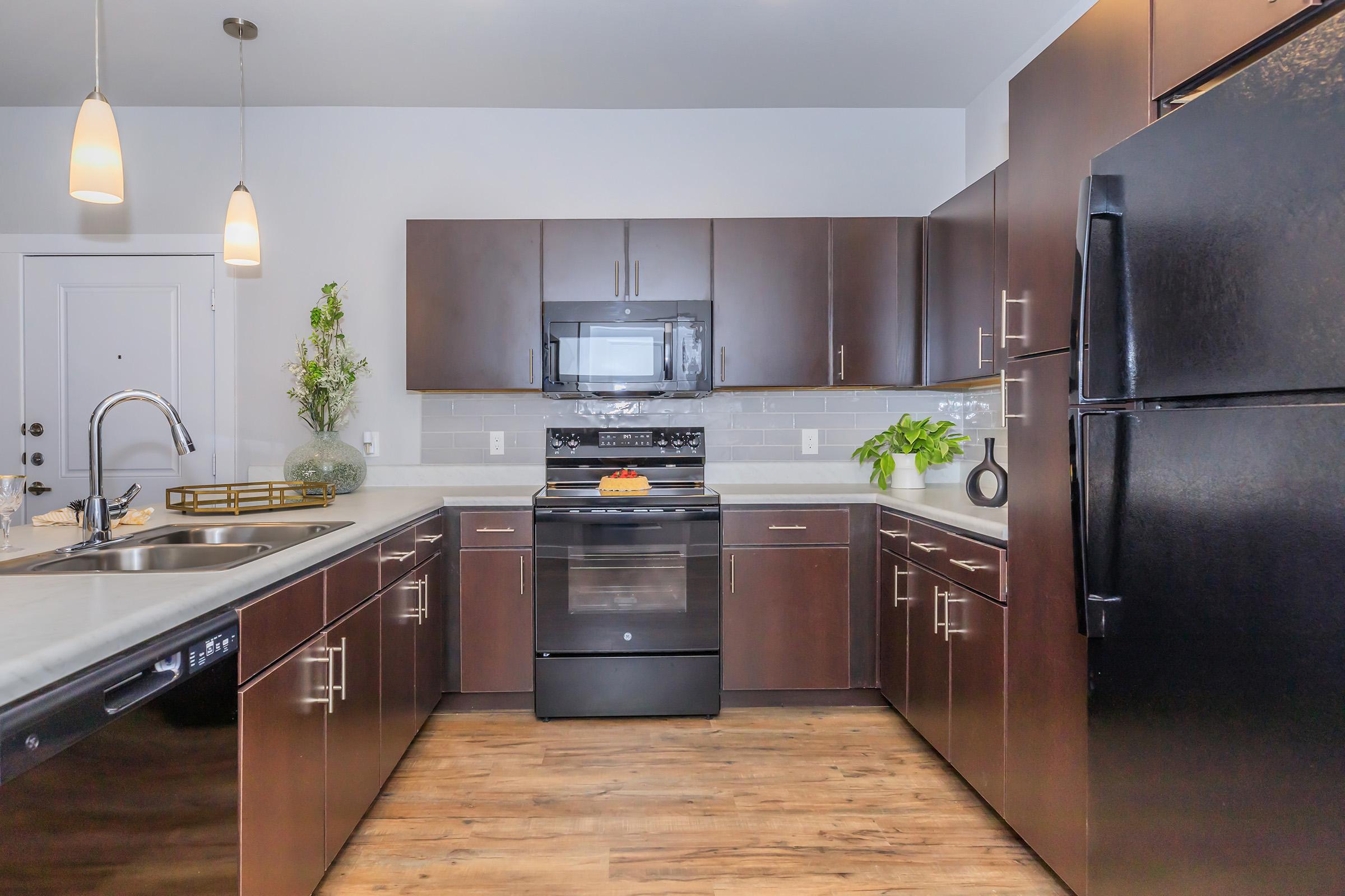 A modern kitchen featuring dark wood cabinetry, stainless steel appliances, and a central island. The countertop includes a stove, microwave, and a sink with a faucet. A black refrigerator stands to the right, while decorative plants and a fruit bowl add color to the space. Pendant lights hang above the island.