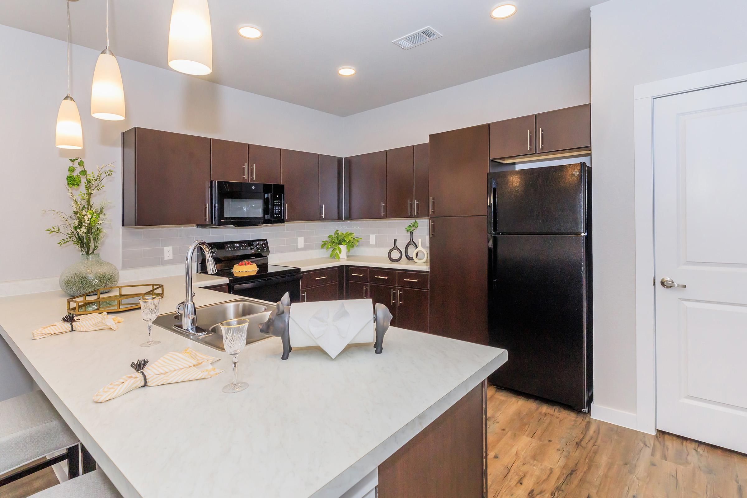 Modern kitchen featuring dark wood cabinets, stainless steel appliances, a black refrigerator, and a white countertop. The space includes pendant lighting, decorative plants, and a dining setup with elegant tableware, creating a stylish and inviting atmosphere.