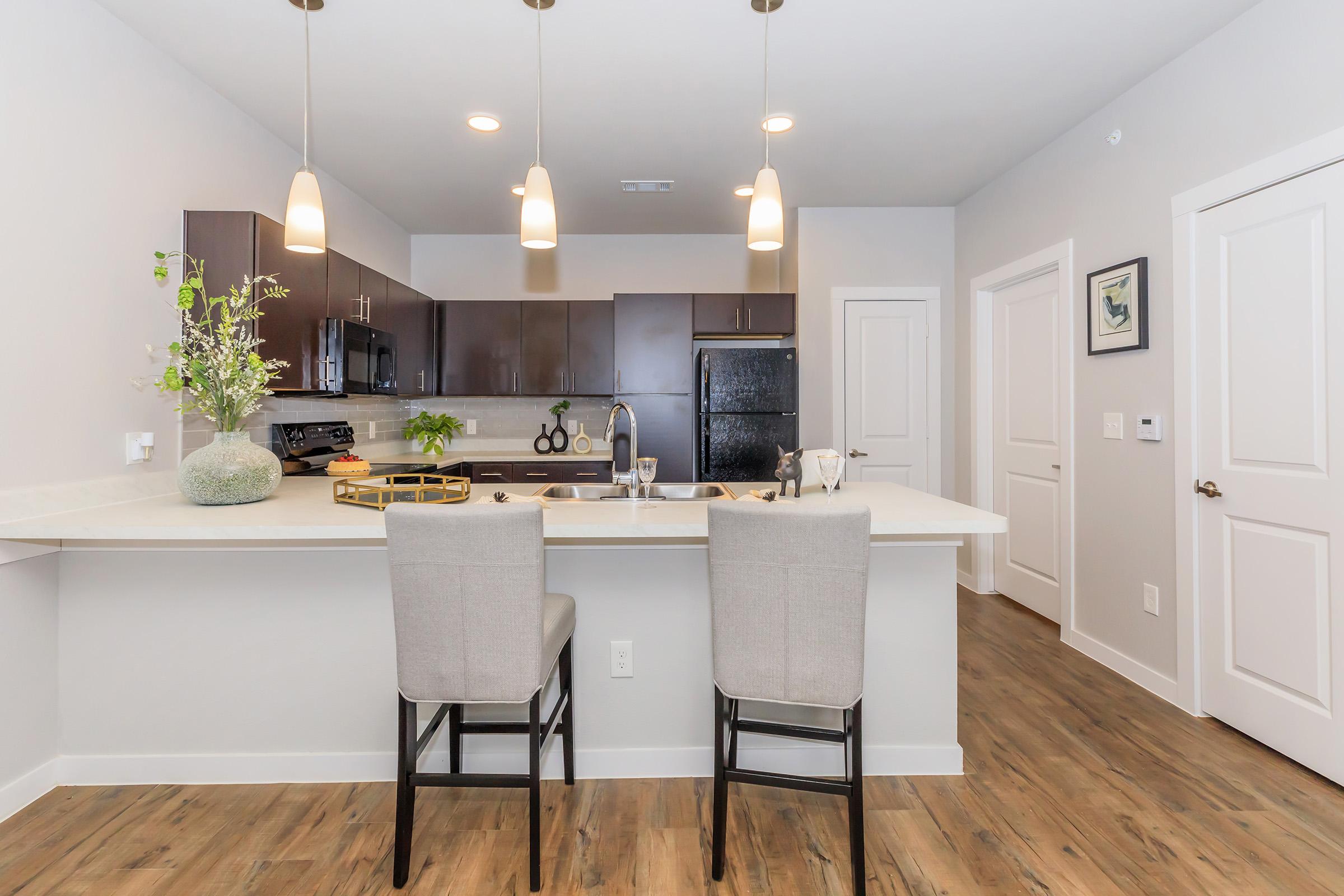 Modern kitchen interior featuring dark wood cabinetry, light countertops, and two barstools at a breakfast bar. Pendant lights hang above the counter, and a black refrigerator is visible. The space has a neutral color palette with a plant arrangement for decoration. Interior doors and a framed picture add to the decor.