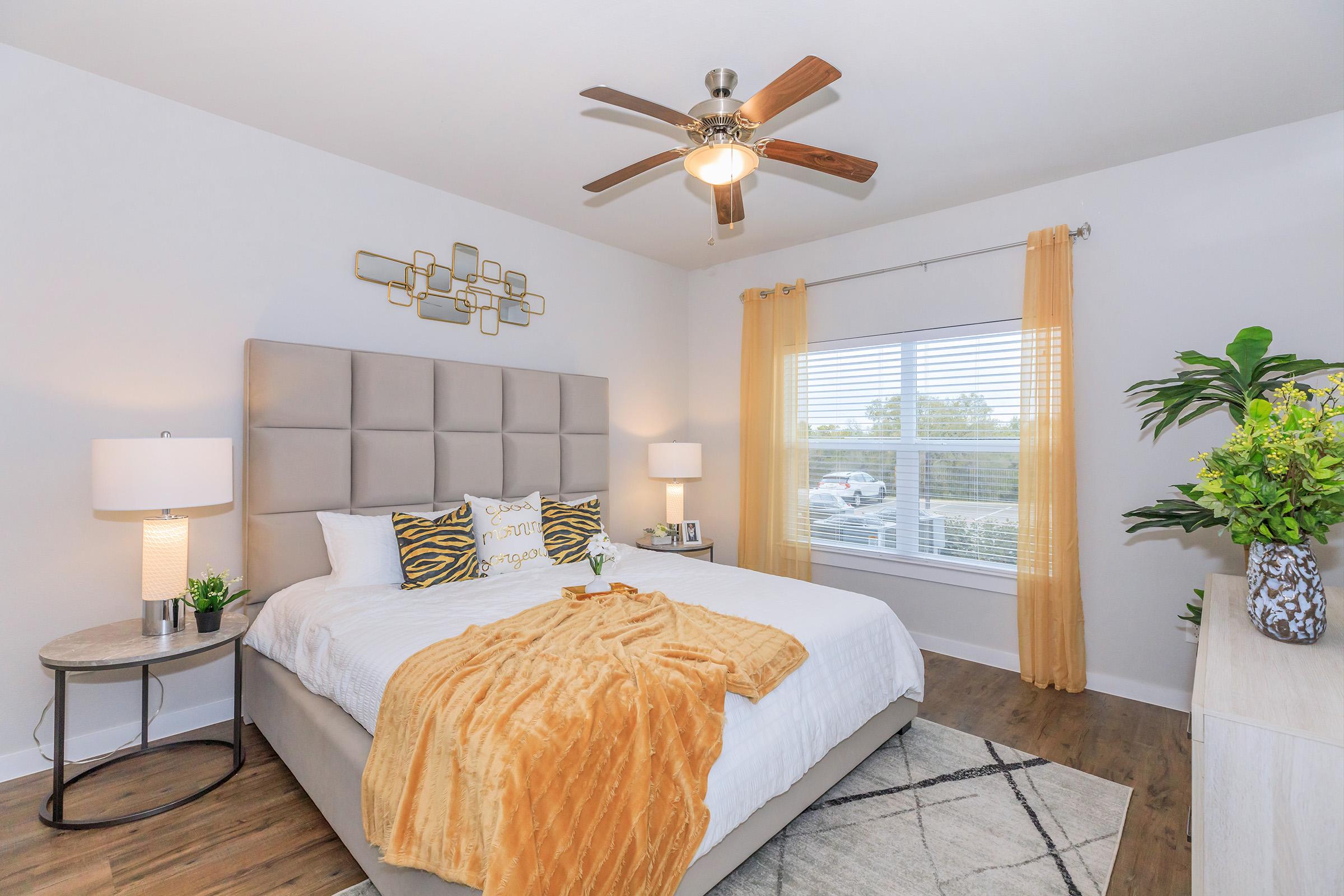 A beautifully decorated bedroom featuring a large bed with a plush headboard, colorful pillows, and a cozy throw blanket. The room has warm yellow curtains, a ceiling fan, and a nightstand with a lamp. A window allows natural light in, complemented by potted greenery and a stylish dresser.