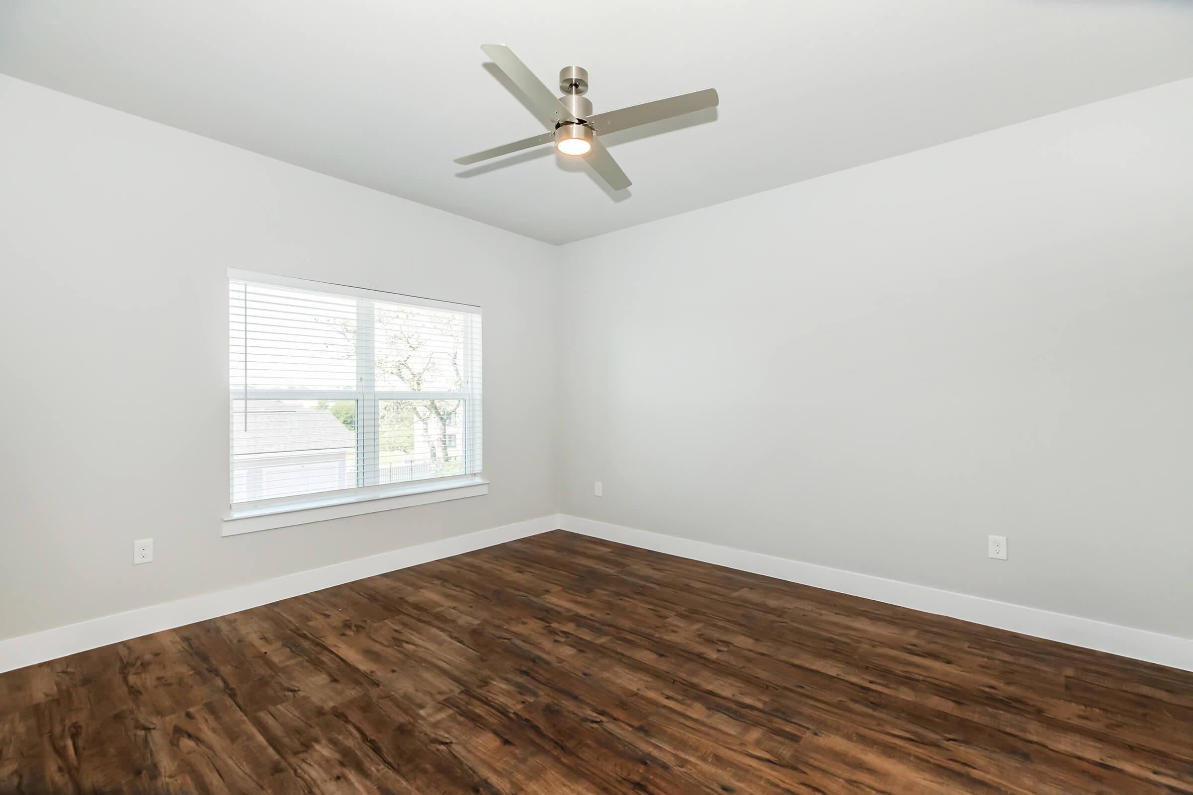 A spacious, empty room with light gray walls and a ceiling fan. Natural light streams in from a window with white blinds, illuminating the dark wooden floor. The room has minimal furnishings, creating a bright and airy atmosphere, ideal for personalization.
