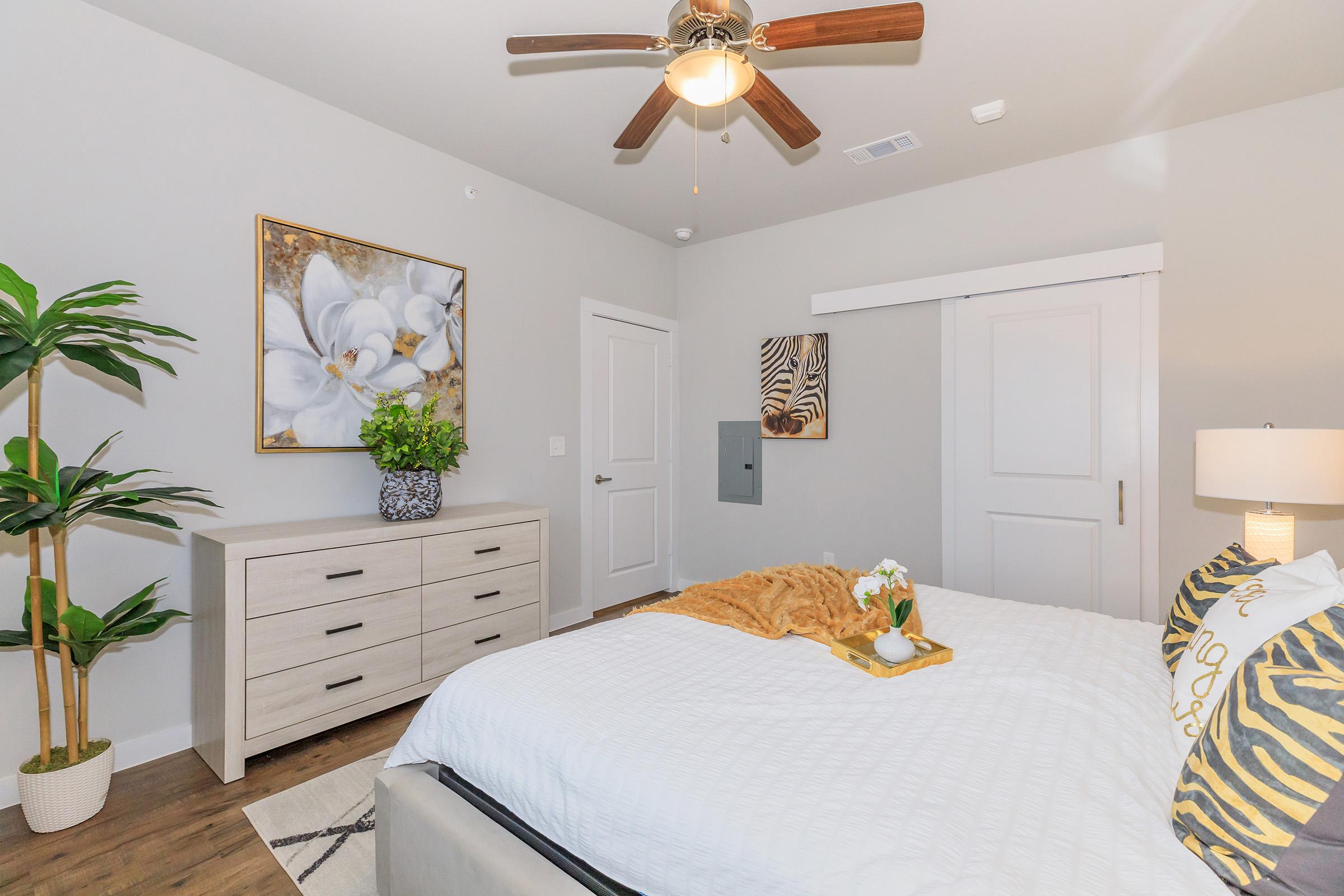 A well-decorated bedroom featuring a light gray color scheme. There is a bed with decorative pillows and a cozy throw blanket. A nightstand with a lamp sits beside the bed. A potted plant and a piece of wall art featuring flowers are in the room, along with a dresser and a sliding door.
