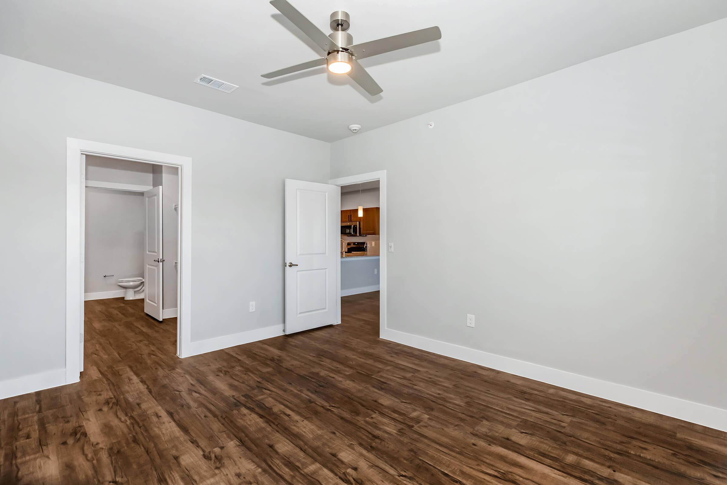 A spacious, modern room featuring light gray walls, a ceiling fan, and hardwood-style flooring. There are two doorways: one leading to a bathroom with a toilet visible, and another leading to a kitchen area. Natural light illuminates the space, creating a bright and inviting atmosphere.