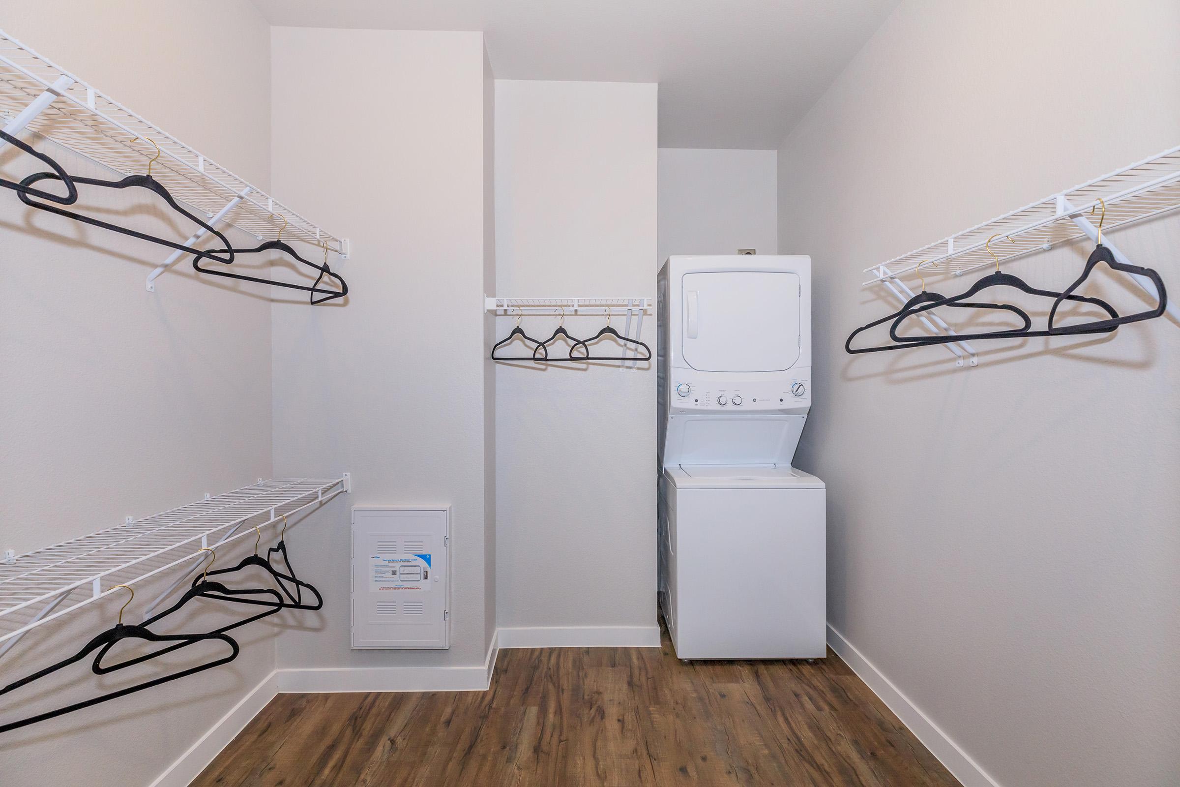 A clean, modern laundry room featuring a stacked washer and dryer unit. The space has white wire shelving along the walls, with several black hangers hanging from the shelves. The floor is made of light-colored wood, and the walls are painted a soft white, creating an airy feel.