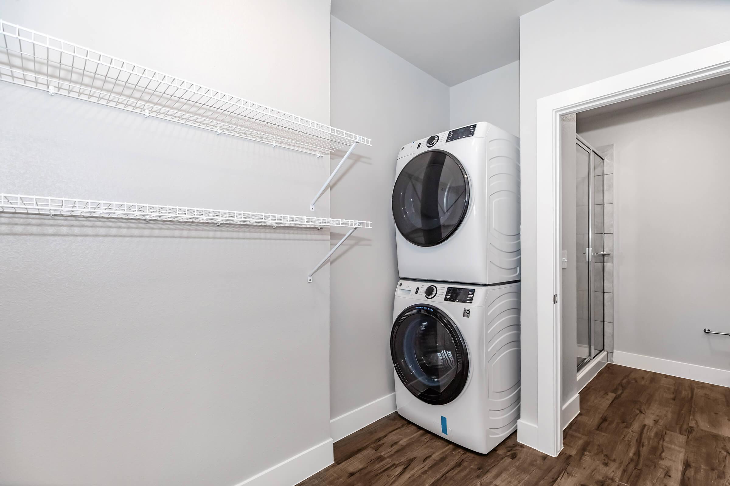 A modern laundry room featuring a stacked washer and dryer in white, along with a shelf for storage. The walls are painted light gray, and the floor is a wood-like laminate. A shower is visible in the background, indicating a clean and functional space.