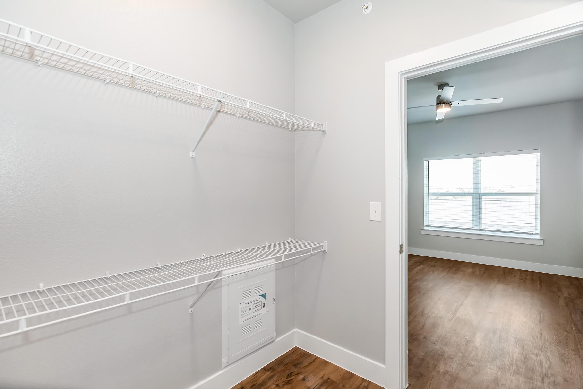 A spacious closet with wire shelving on one side and a door leading to a well-lit room featuring a window with blinds and a ceiling fan. The walls are painted light gray and the floor is made of wood.