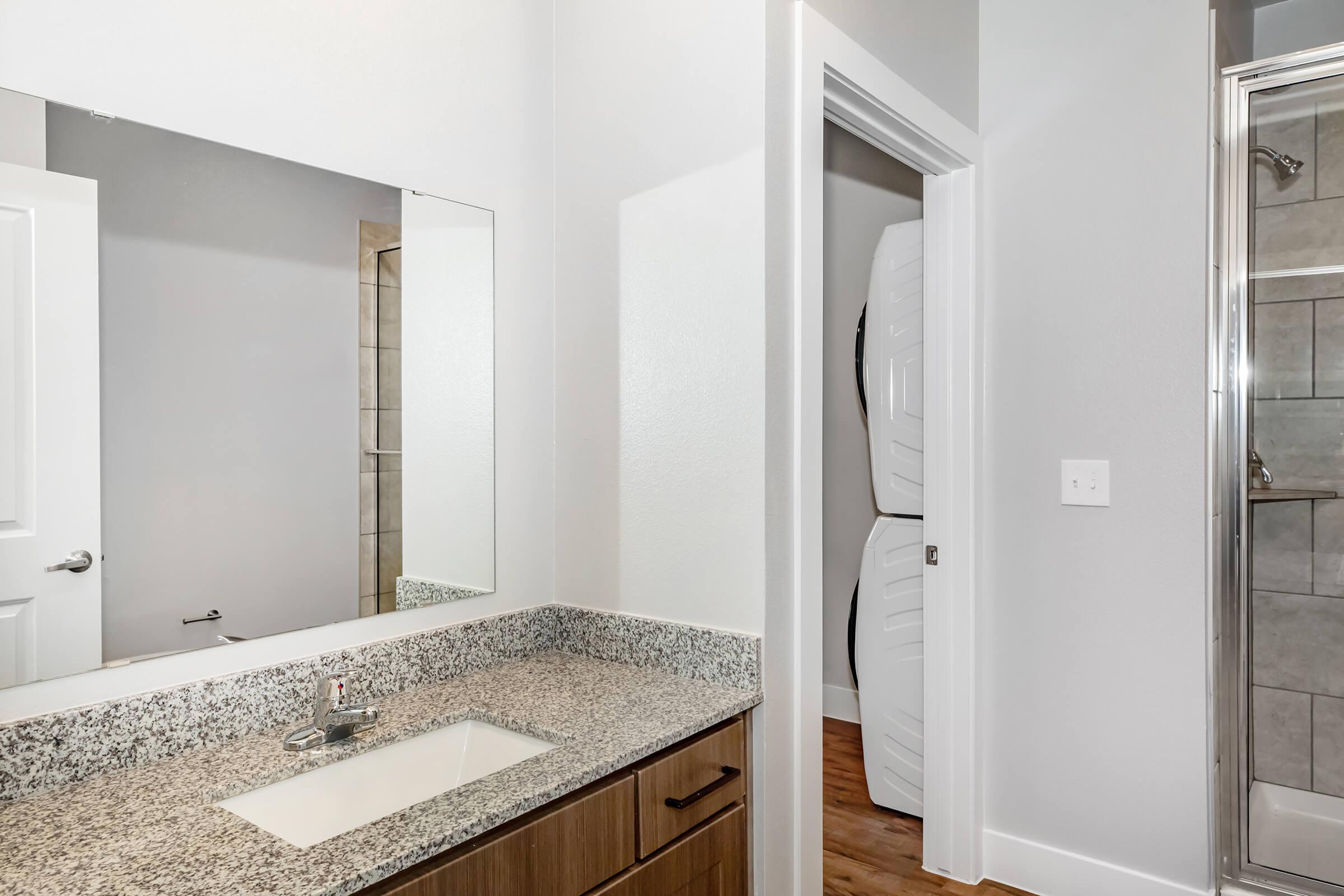Modern bathroom featuring a granite countertop with a sink, a large mirror, and a view of an adjacent shower. The decor includes light-colored walls and wood flooring. A door leads to another room, and a stack of materials is visible nearby.