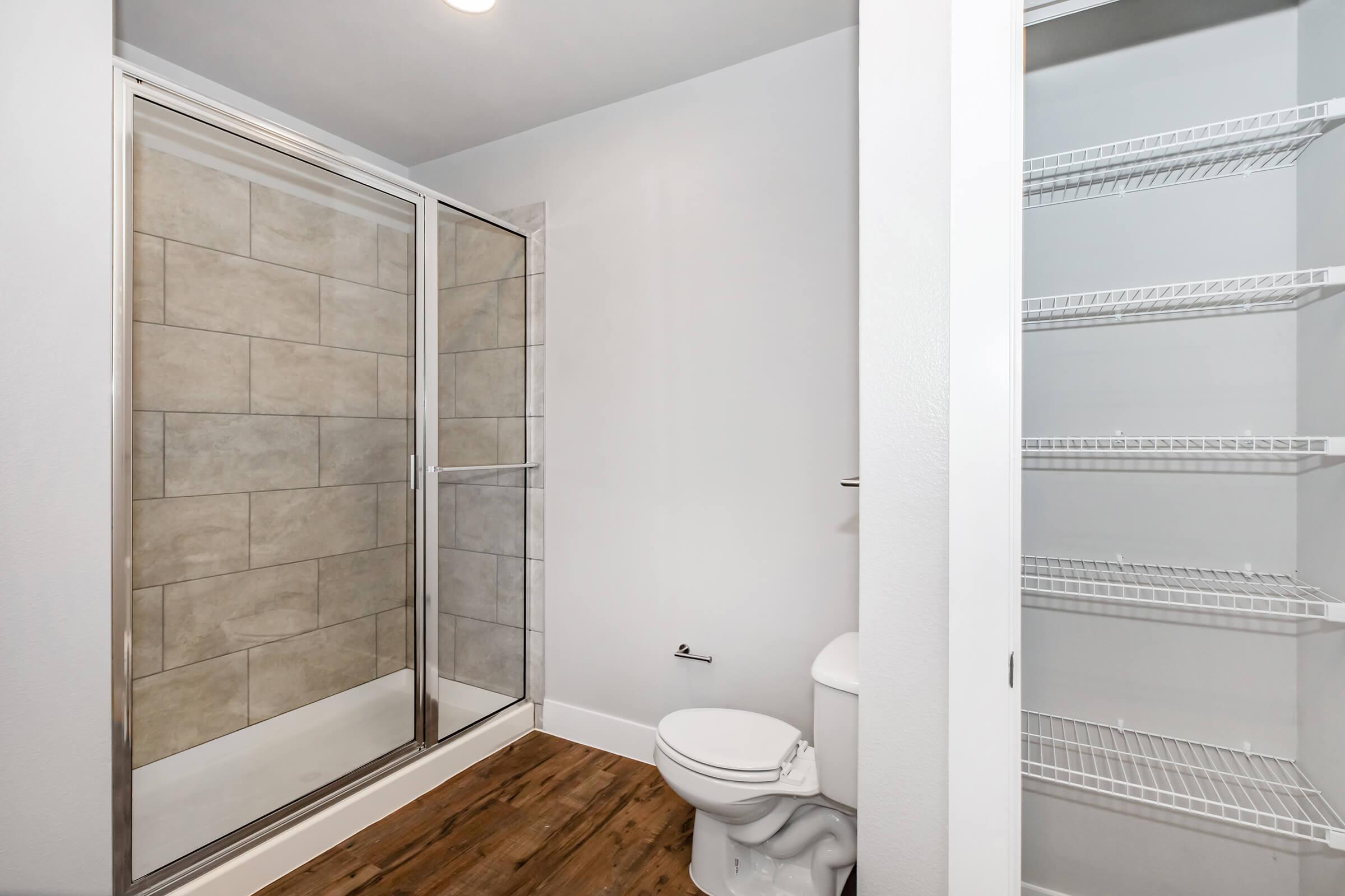 A modern bathroom featuring a glass shower enclosure, a white toilet, and light-colored tiled walls. The floor is wooden, and there is a white shelving unit on the right side, providing storage space. The walls are painted in a soft, neutral color.