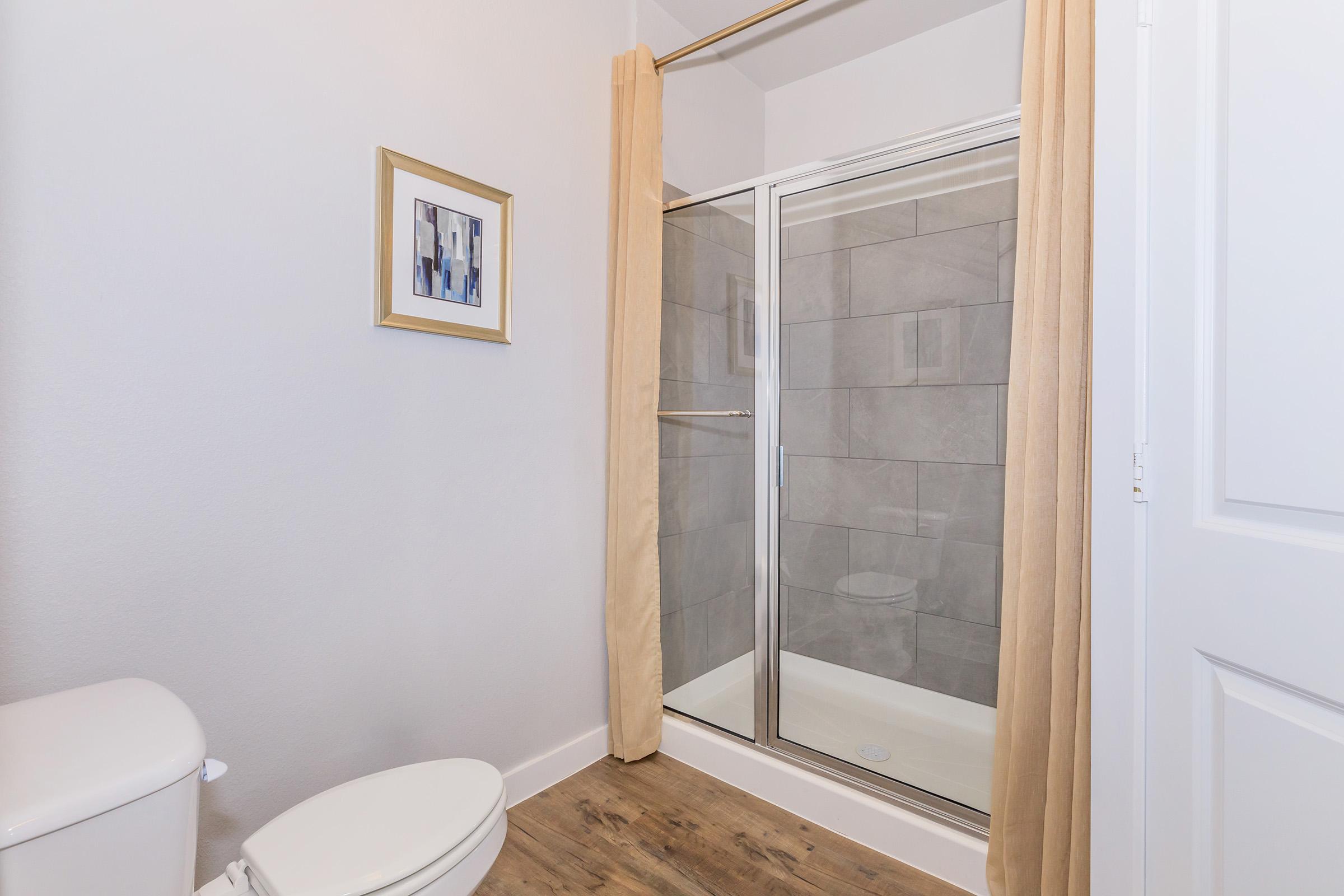 A clean and modern bathroom featuring a white toilet, a glass shower enclosure with beige curtains, and light gray tiled walls. A framed abstract artwork hangs on the wall, complementing the neutral color scheme and wooden flooring.
