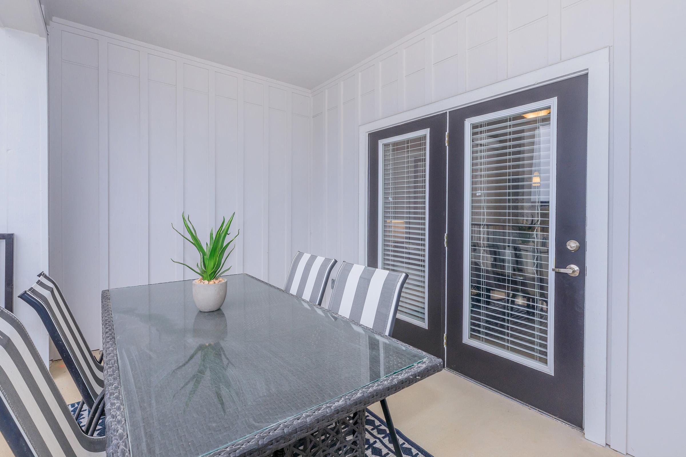 A modern dining area featuring a glass-top table with black and white striped chairs. A potted plant sits at the center of the table. There are double doors with vertical blinds leading to another space, surrounded by white paneling on the walls. The floor is light-colored and minimalistic.