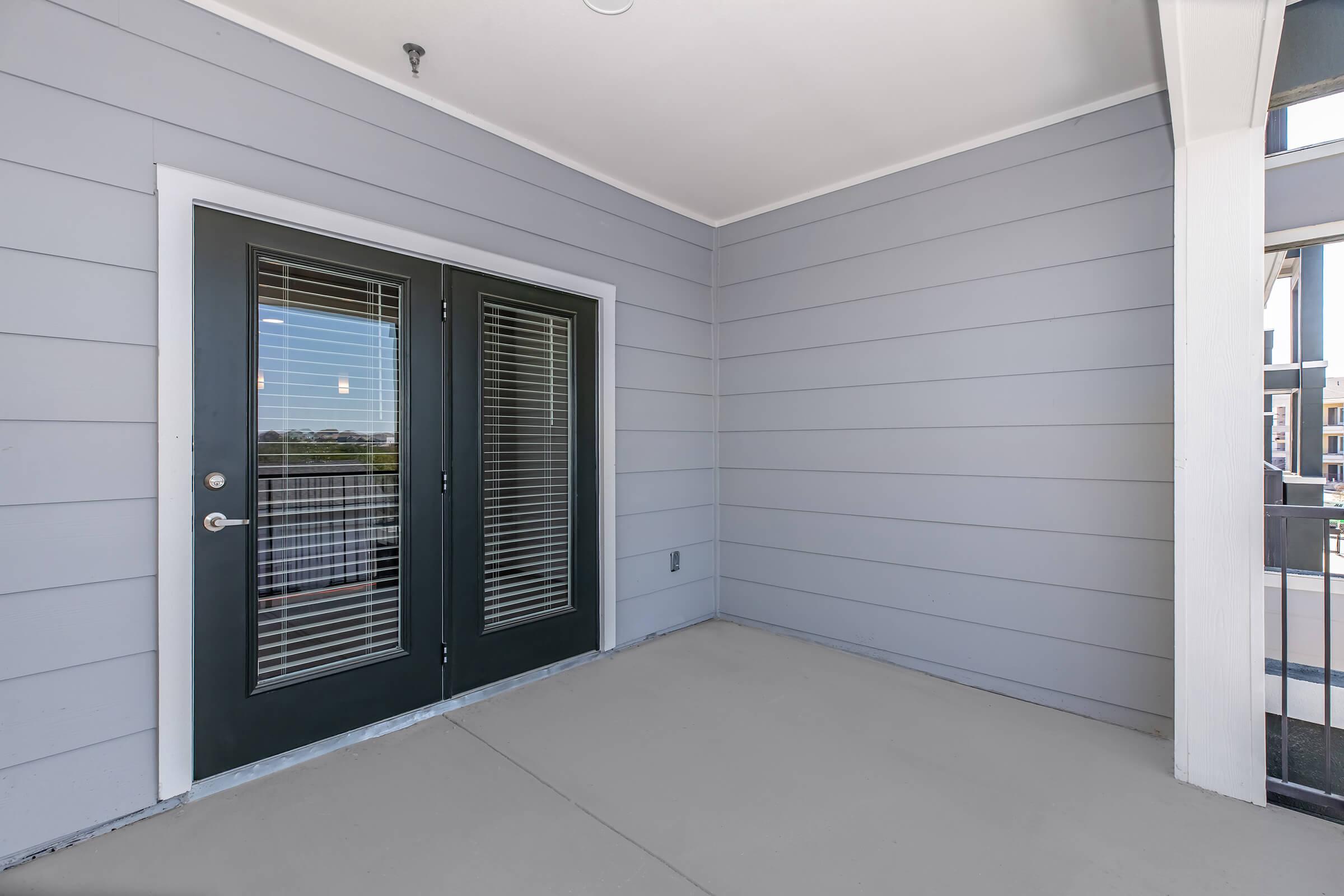 A small outdoor balcony area with gray siding walls, featuring double glass doors with dark frames and vertical blinds. The floor is a light concrete color, and there is a railing visible in the background, indicating a second-floor location. The area is uncluttered and well-lit.