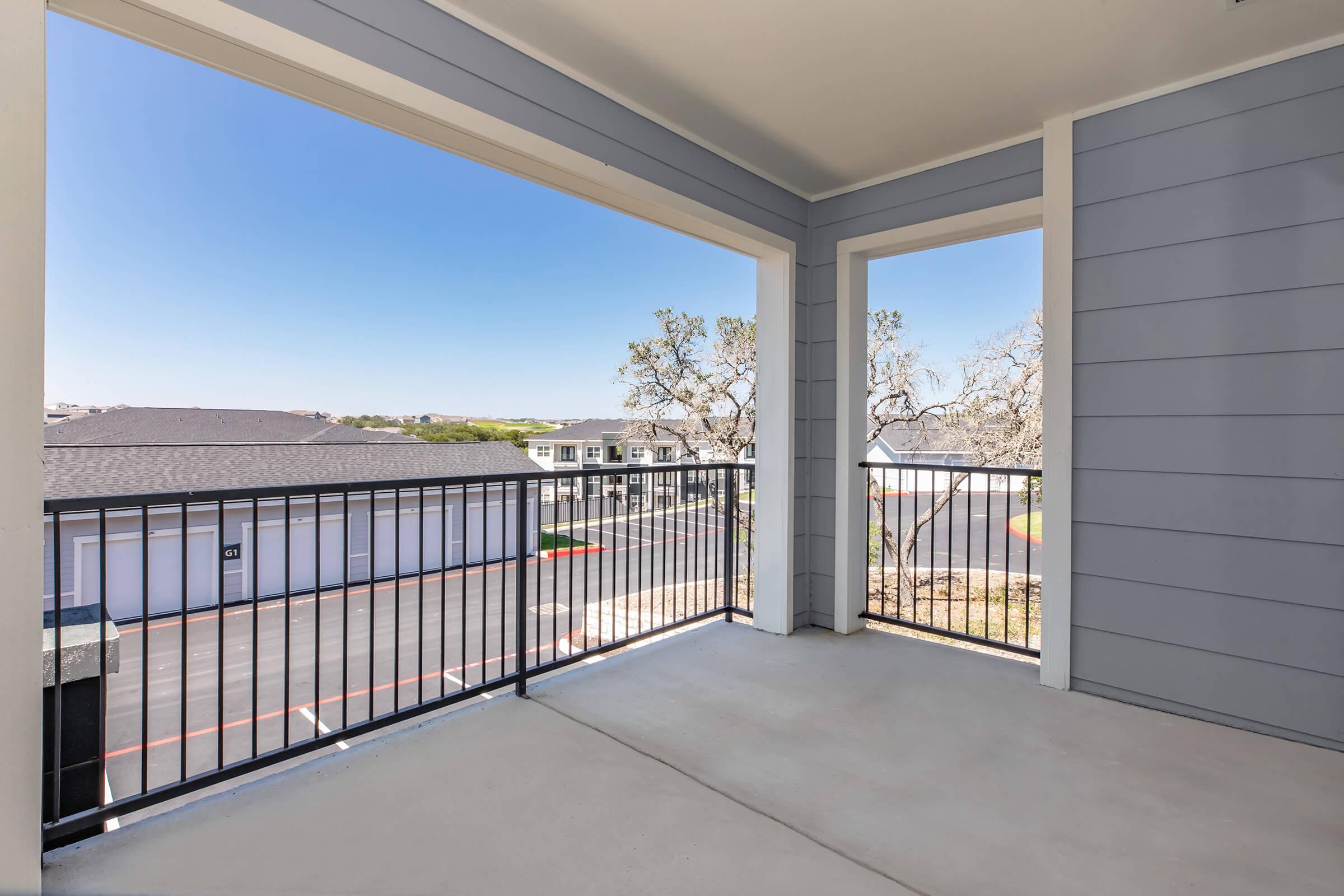 A spacious balcony with a black metal railing, offering a view of the surrounding area, including buildings and trees. The clear blue sky adds brightness to the scene, creating a pleasant outdoor space for relaxation. The floor is concrete, and the walls are painted in light gray.