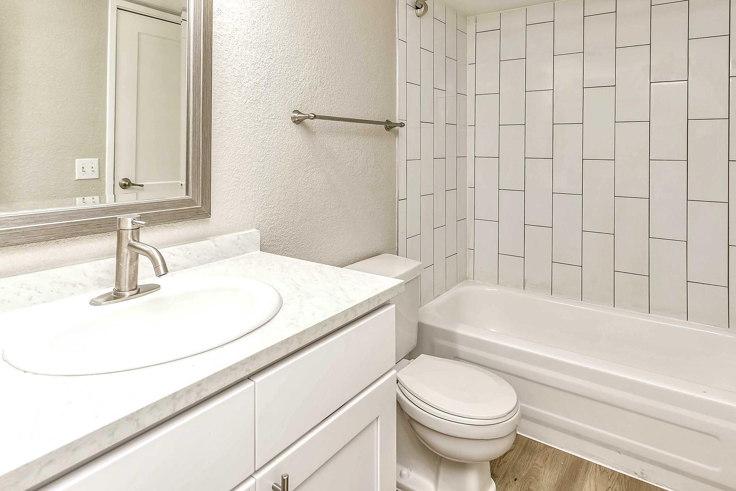 A modern bathroom featuring a white bathtub, a sink with a marble countertop, and a mirror above the sink. The walls are tiled in a light color with vertical white tiles. A towel bar is mounted on the wall, and the floor has a wooden finish, creating a clean and contemporary look.