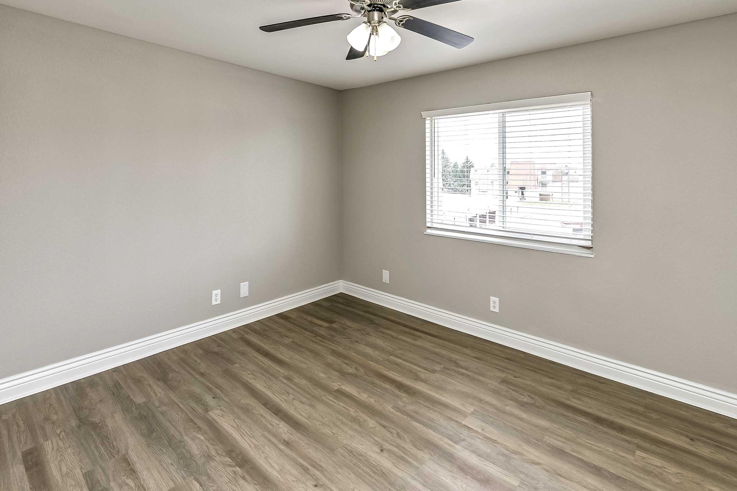 Empty room featuring a light-colored wall, a ceiling fan, and a window with blinds allowing natural light. The flooring is made of laminate with a wooden appearance. There are no furnishings or decorations, creating a spacious and minimalist atmosphere.