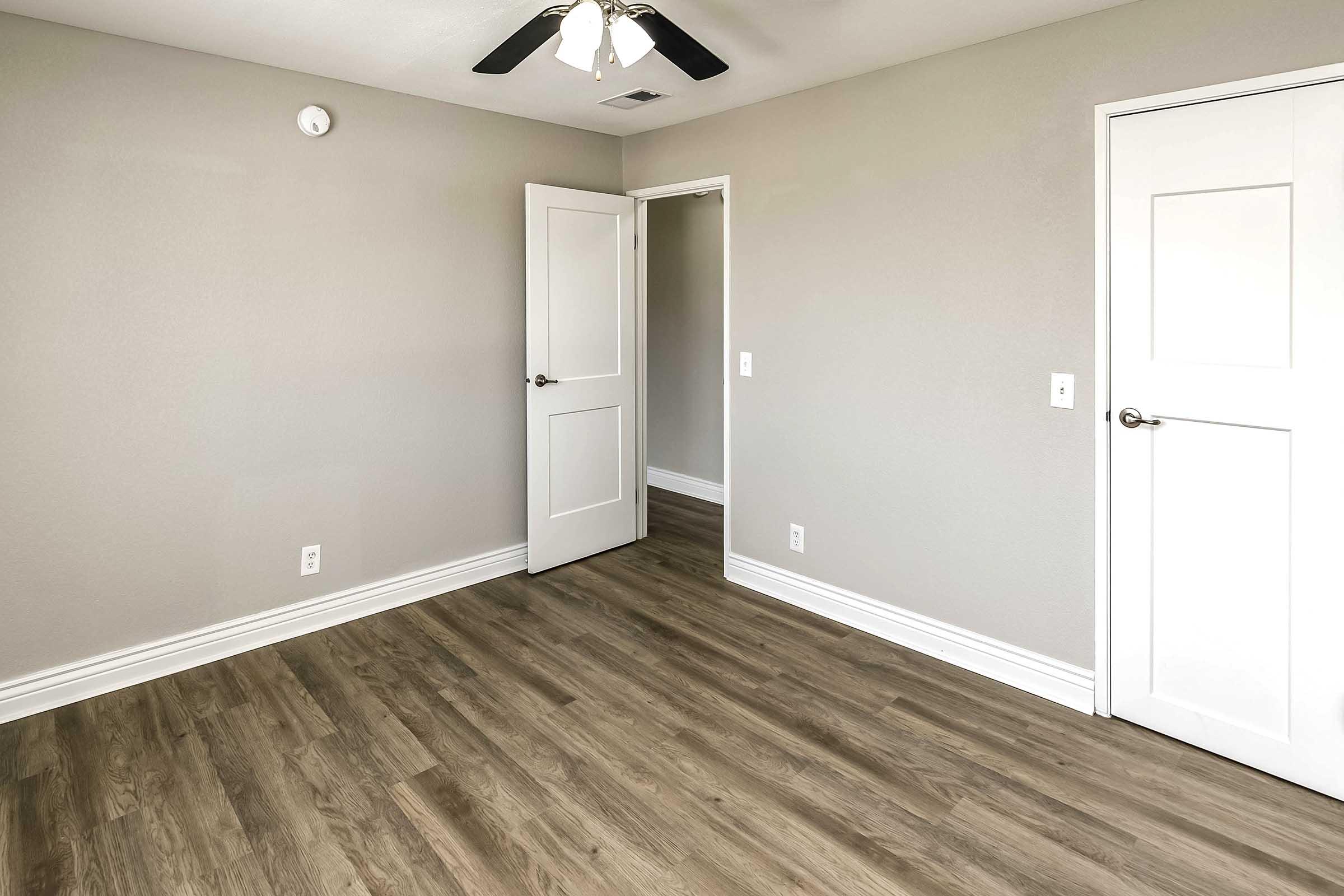A minimalistic empty room with light gray walls and a wooden floor. It features a ceiling fan, a closed door, and an open door leading to another small space. The room is well-lit, and there are no furnishings, creating a spacious and clean atmosphere.