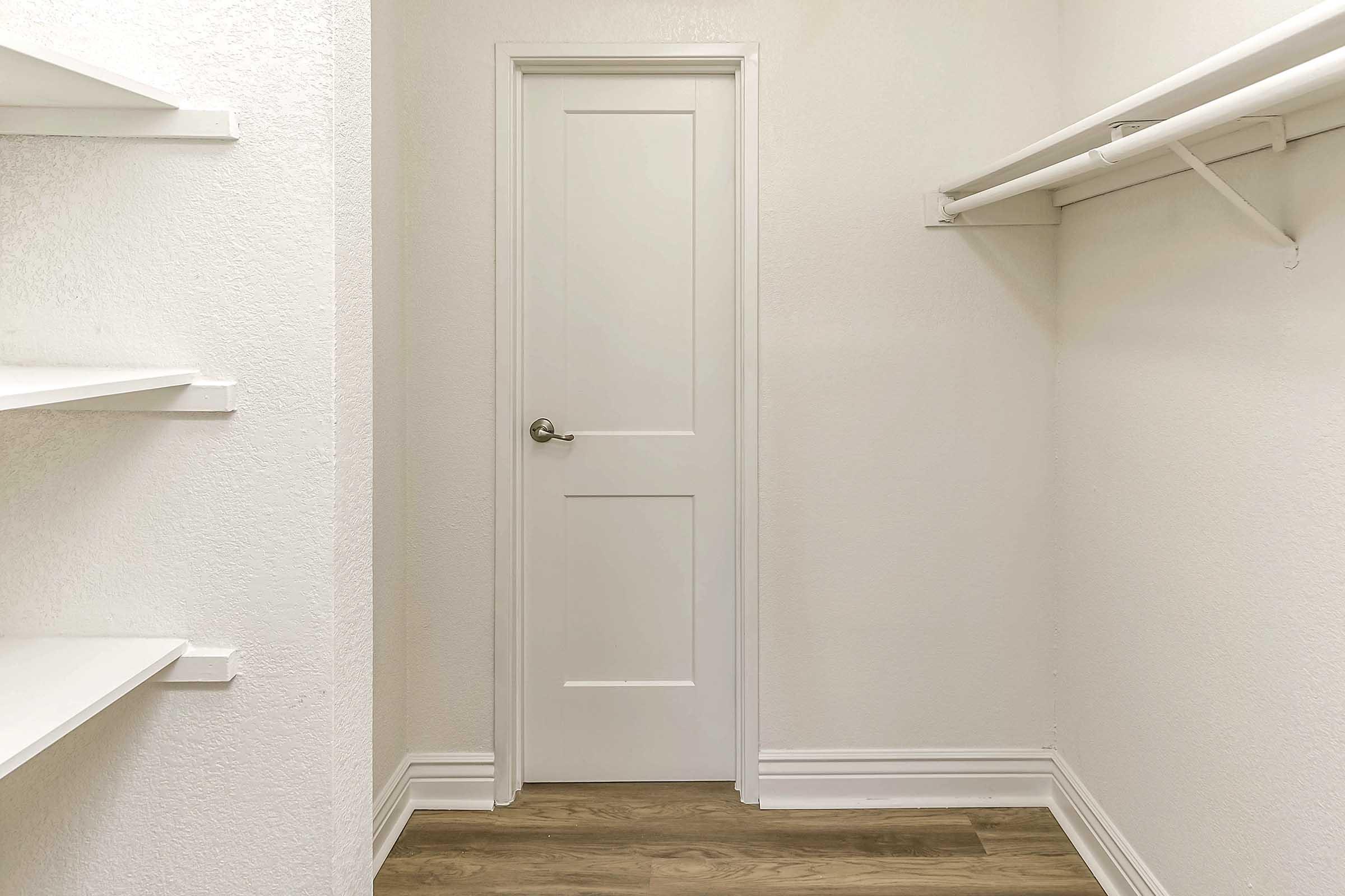 A small, empty room with white walls and a light wooden floor. It features a closed white door and two shelves on the left side. The space appears to be a closet or storage area, well-lit and tidy, with ample room for organizing items.