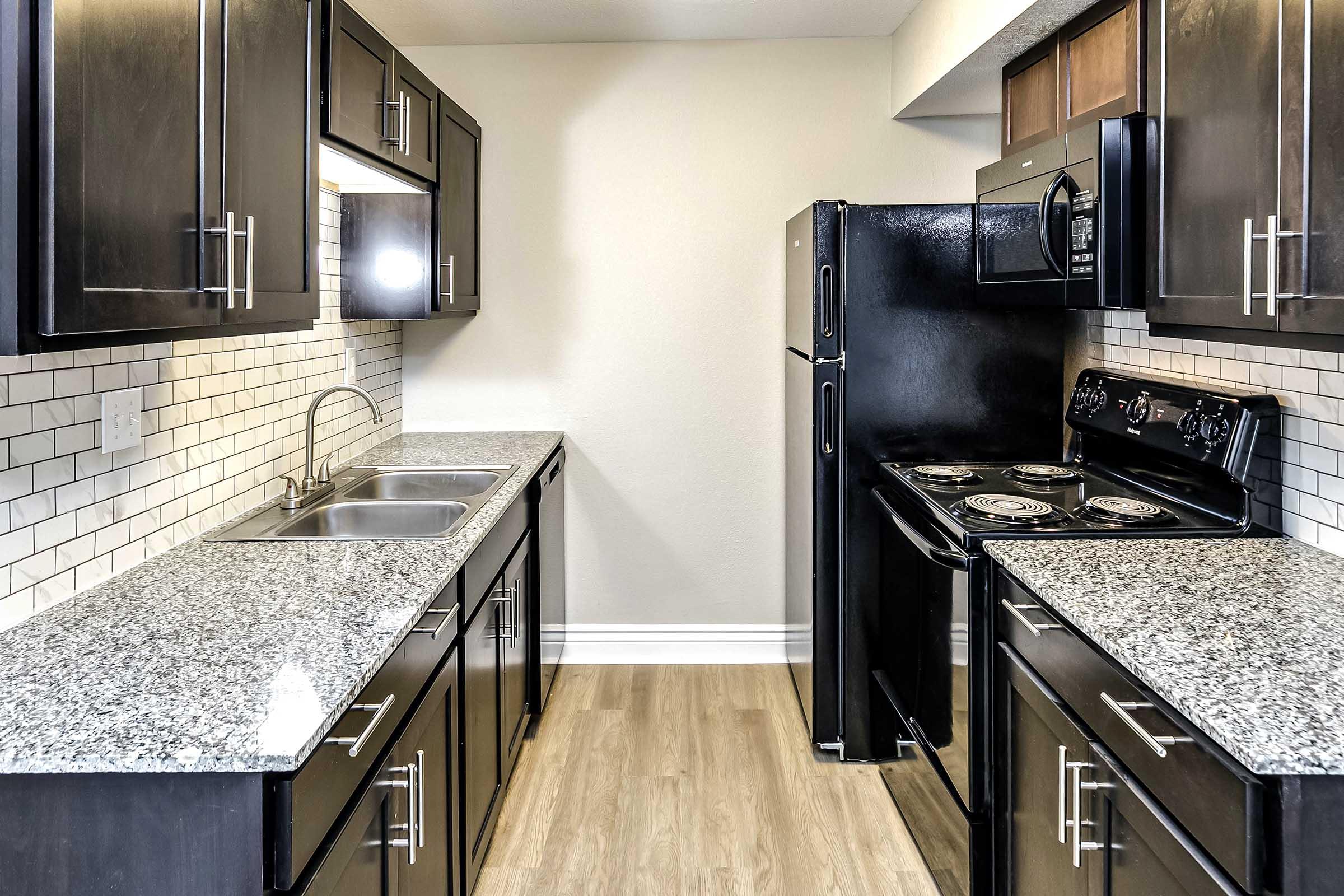 Modern kitchen featuring dark cabinetry, granite countertops, stainless steel double sink, and black appliances including a refrigerator and stove. The backsplash is designed with white subway tiles, and the flooring is light wood, creating a bright and inviting atmosphere.