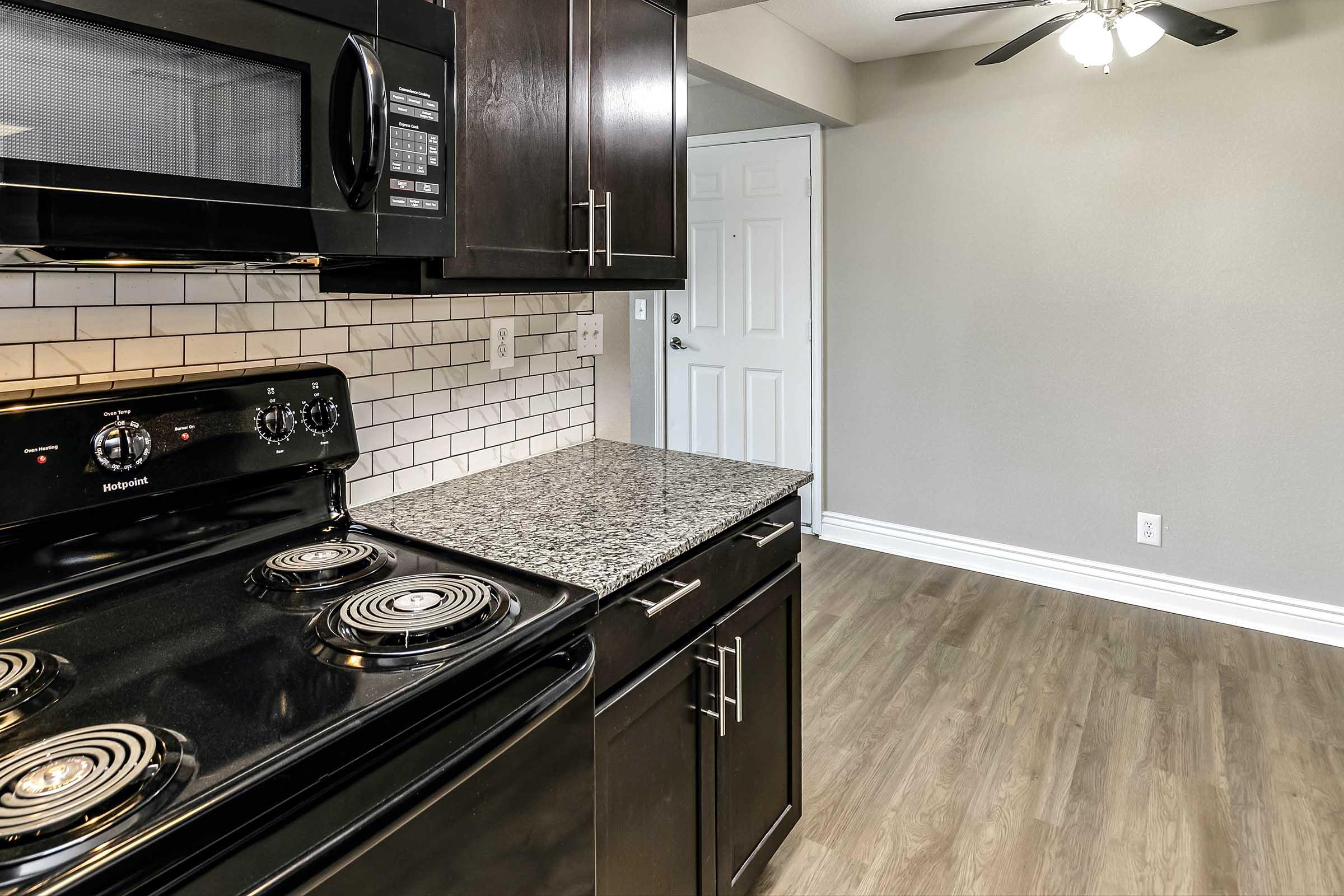 A modern kitchen featuring a black stove with four burners, a microwave above, and dark wood cabinets. The countertop is made of speckled gray stone. The background shows a wall with a door and a ceiling fan, all set against a light gray wall with laminate flooring.