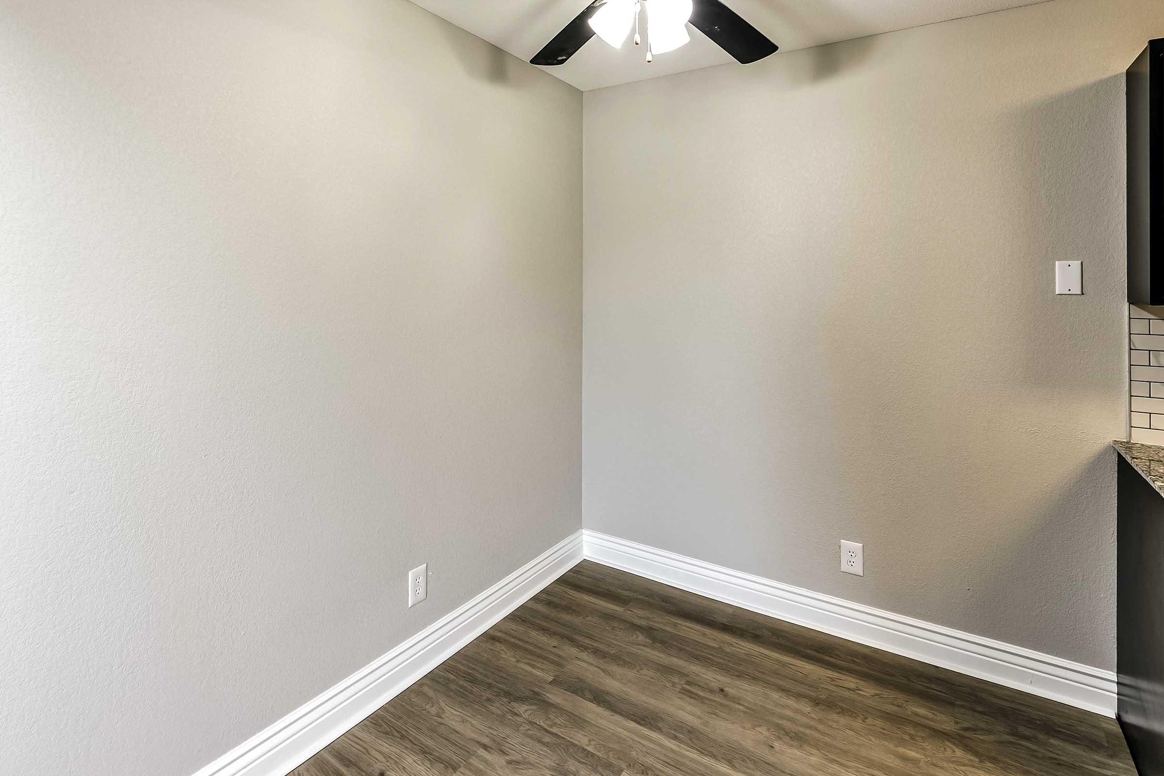 An empty room with light gray walls and a ceiling fan. The floor is covered with wooden laminate. There is a small area with dark cabinetry, and the overall space is well-lit with natural light. The room has a clean and modern appearance, suitable for various uses.
