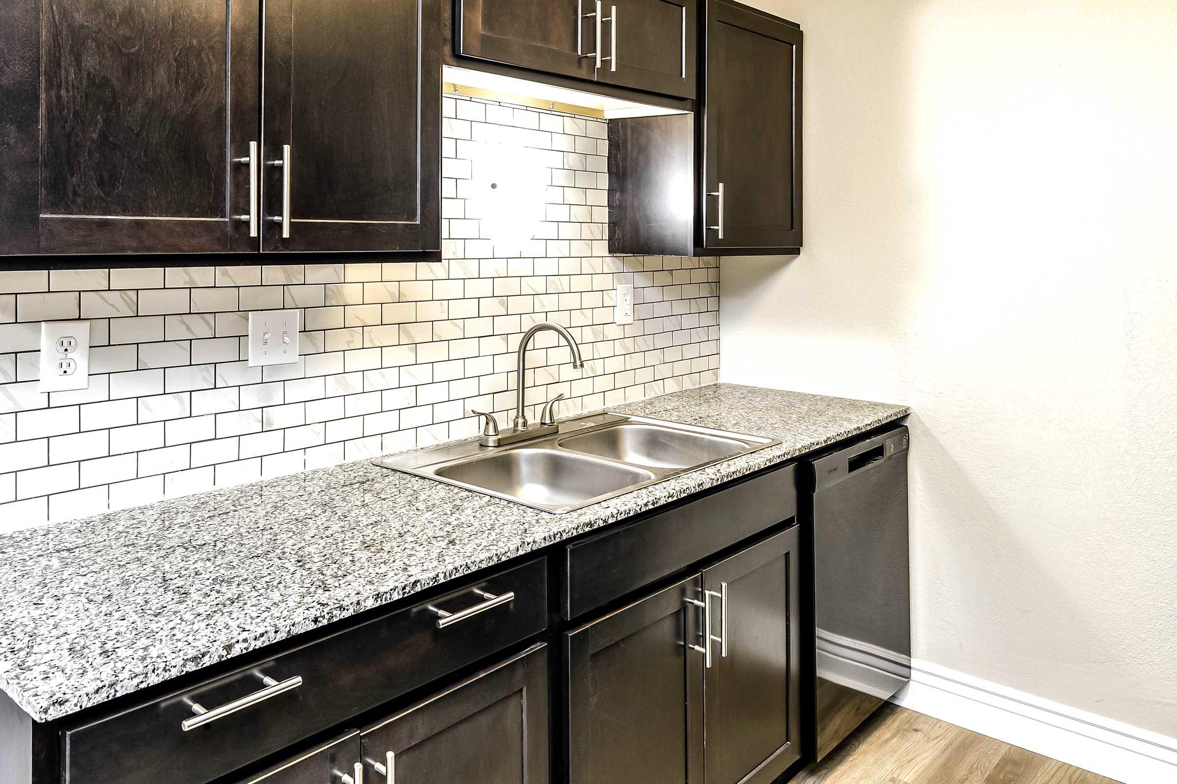 Modern kitchen with dark wood cabinets, a double sink, and granite countertops. White subway tile backsplash enhances the design. Appliances include a dishwasher, with countertop space extending on both sides. The overall color scheme is neutral and contemporary.