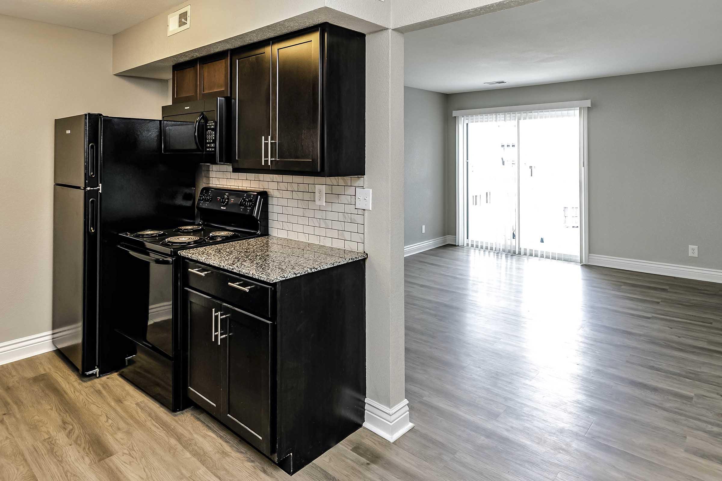 Modern kitchen featuring black appliances, including a refrigerator and stove, alongside dark cabinetry and granite countertops. An open layout leads to a bright living area with a sliding glass door that opens to a balcony or outdoor space. The flooring is a light-colored laminate.